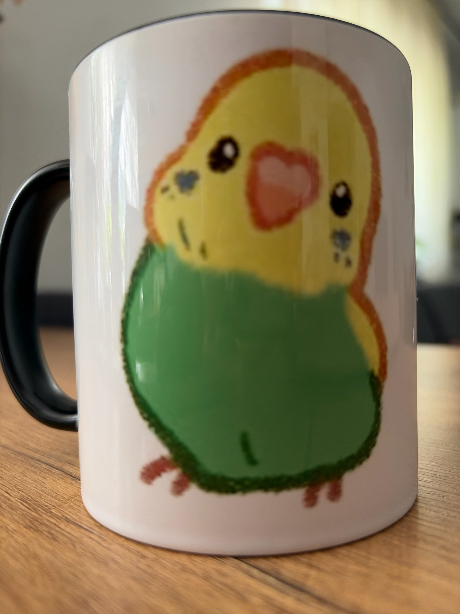 A close-up shot shows a white ceramic mug with a black handle. The mug features a cartoon illustration of a yellow and green budgie. The budgie has a pink heart-shaped beak, black eyes, and red feet. The mug is placed on a wooden surface, likely a table. The background is out of focus, with hints of a room and natural light. The scene is set in Oxentea, Moldova, indicating a domestic setting.