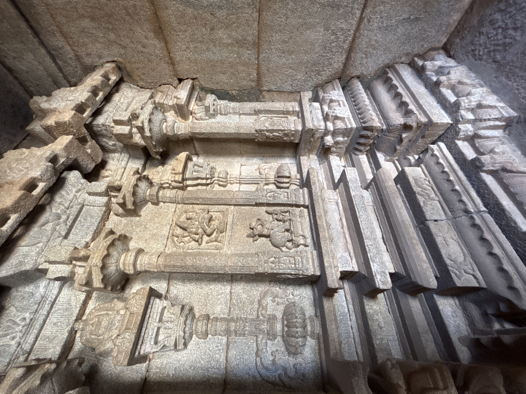 This image depicts the interior stonework of the Hazara Rama Temple, located in Kamalapura, India. The scene is an upward-angled view of sculpted walls and ceiling, showcasing detailed carvings in a light grey stone. The walls feature horizontal beams with decorative column-like structures. Carved depictions of figures and animals are integrated into the stonework. The architectural elements have a rough, aged texture. The ceiling is composed of plain stone blocks. Lighting within the scene casts shadows, highlighting the intricate details of the carvings.