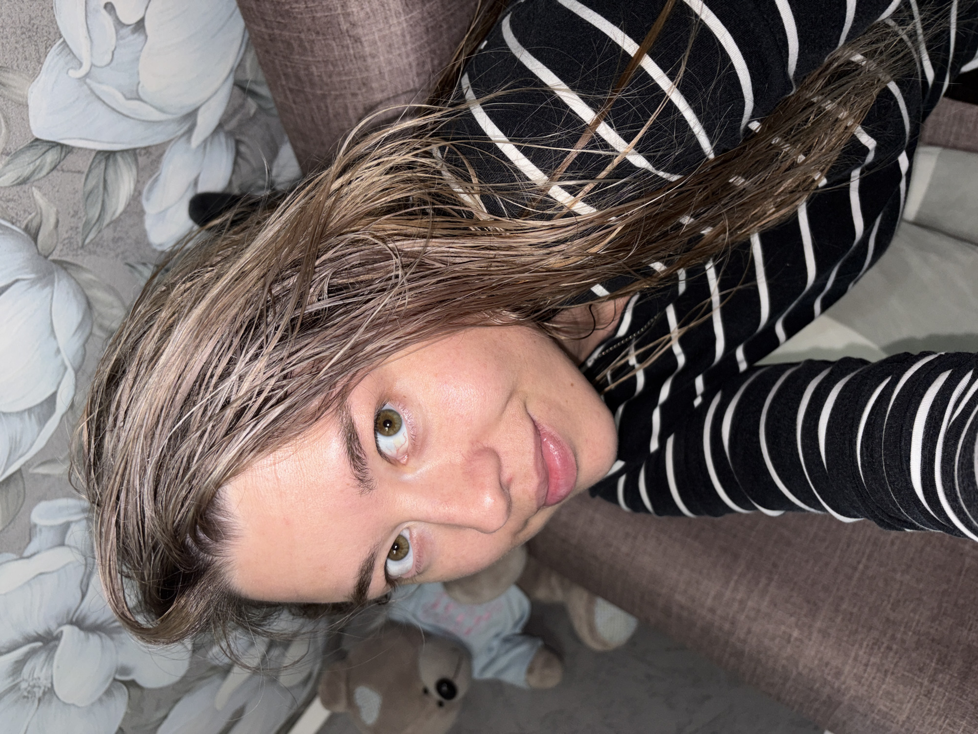 The image is an indoor, eye-level selfie featuring a young woman looking directly into the camera with a subtle, calm expression. Her hair, which appears damp and light brown or blonde, frames her face. She has prominent green or hazel eyes and slightly parted lips. She is dressed in a long-sleeved top with a black and white horizontal stripe pattern.
The setting appears to be a casual domestic environment, likely a living room or bedroom. To her left and behind her, the wall is covered with a patterned wallpaper featuring large, stylized white and light grey floral designs. A textured brown upholstered piece of furniture, possibly a couch or armchair, is visible on her right and extends into the lower part of the frame.
Below and to the left of the woman's face, a small, light brown teddy bear is partially visible, resting on the brown furniture. The teddy bear is wearing a light blue top with an indistinct light-colored design or text across its chest. The lighting in the room appears to be artificial and somewhat soft, creating an intimate atmosphere. The location context for this scene is Chaikovsky, Russia.