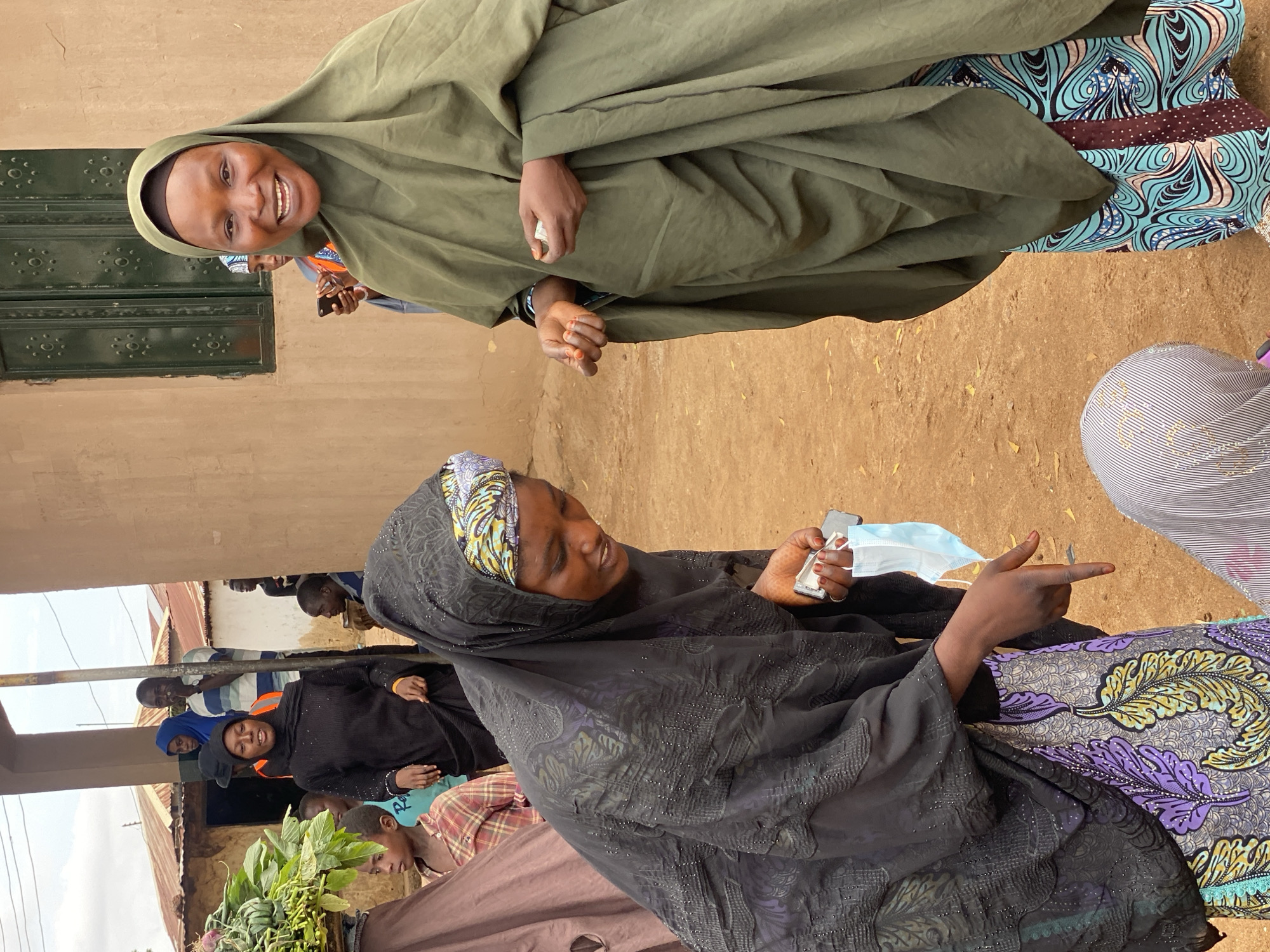 In Rigachikun, Nigeria, a group of individuals, primarily women wearing hijabs, are gathered outdoors. The scene appears to be informal, possibly a community gathering or a moment of social interaction. The ground is dusty, and a building with a green-painted door is visible in the background, suggesting a rural or semi-urban environment.
Two women are prominently featured in the foreground. One, wearing a green hijab, smiles warmly at the camera. Her hand is visible, holding a small white object, possibly a piece of paper or fabric. Next to her, another woman in a black hijab is looking down and to the side, holding a blue face mask and gesturing with her hand. Her expression is thoughtful. In the background, other people, including men and children, can be seen, some engaged in conversation or observing the scene.
The attire of the individuals, particularly the women's hijabs and patterned clothing, suggests the cultural context of the location. The presence of the face mask may indicate ongoing public health concerns or practices. The lighting suggests it is daytime, possibly late morning or afternoon, with natural light illuminating the scene. The overall mood conveyed by the visible expressions is one of community and interaction. There is no visible text in the image.