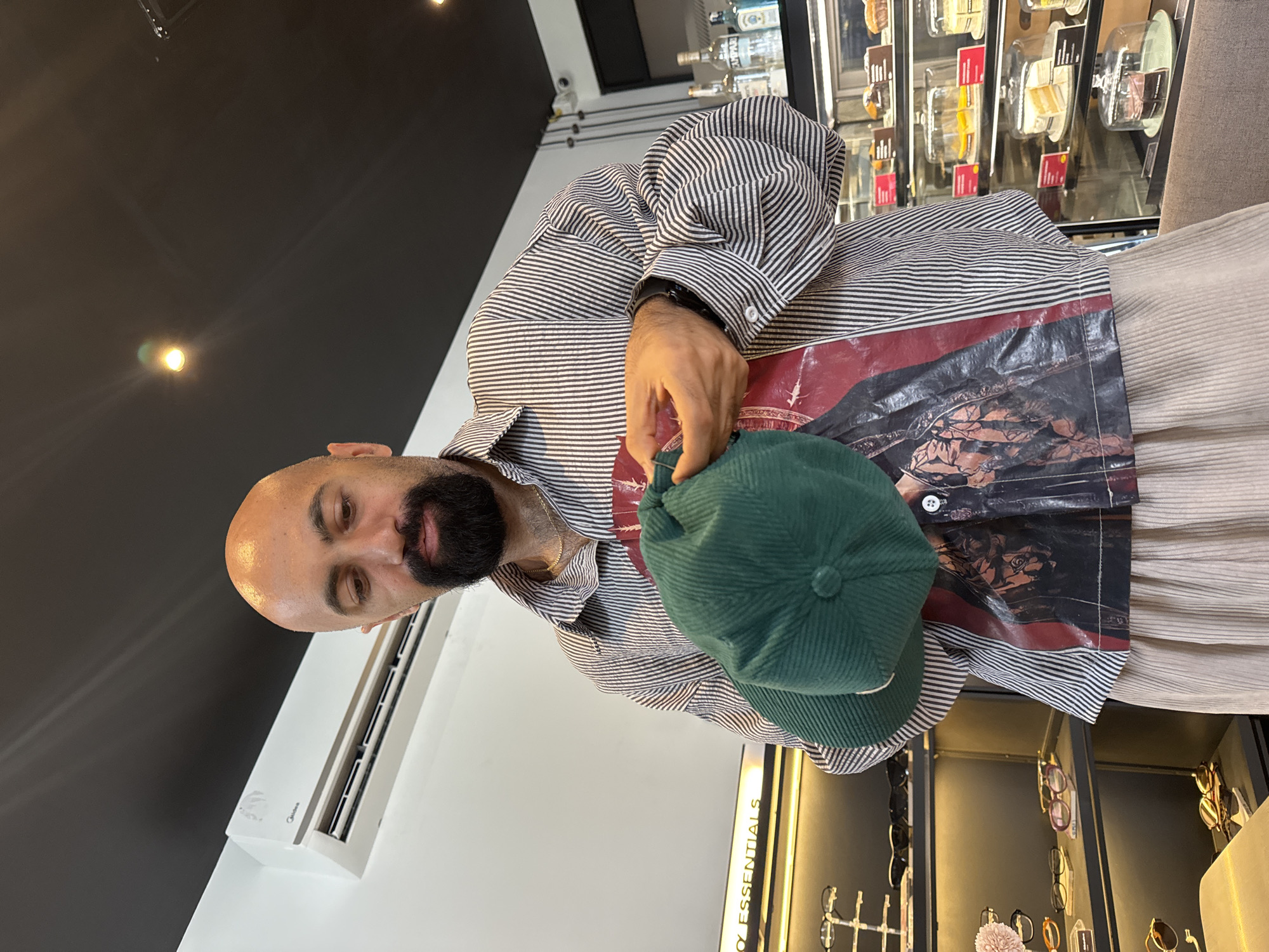 A bald, bearded man wearing a striped shirt, a graphic t-shirt underneath, and light-colored pants holds a green corduroy cap in front of him. The man stands in what appears to be a modern cafe or shop, indicated by display shelves containing various items, including eyewear and pastries. An air conditioning unit is visible on the wall behind him, and a black ceiling with lights is above. The setting suggests a commercial establishment, possibly located in a city like Ko Samui District, Thailand, given the tropical or warm climate implied by the indoor cooling unit.