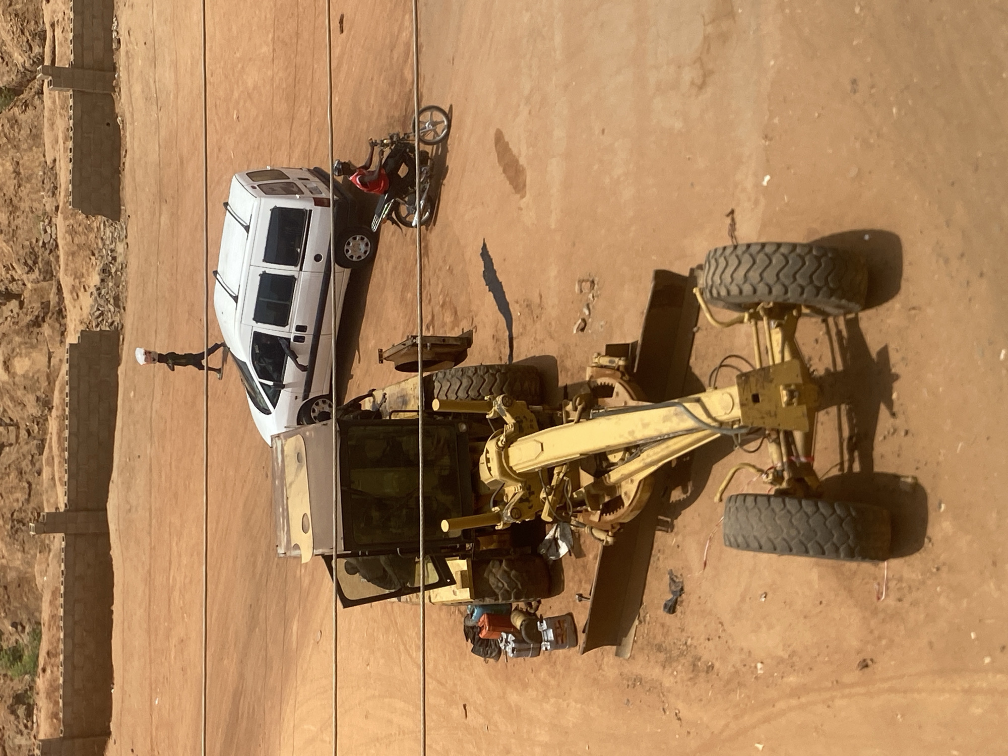 The image, taken from an elevated position in Tudun Wada, Nigeria, shows a construction site or a work area. A white van is parked near a yellow road grader. A motorcycle is parked next to the van. There are two individuals visible: one standing near the van and one seated on the motorcycle. The ground appears to be dirt or sand. There is a wall along one edge of the area. Some tools and equipment are visible near the grader.