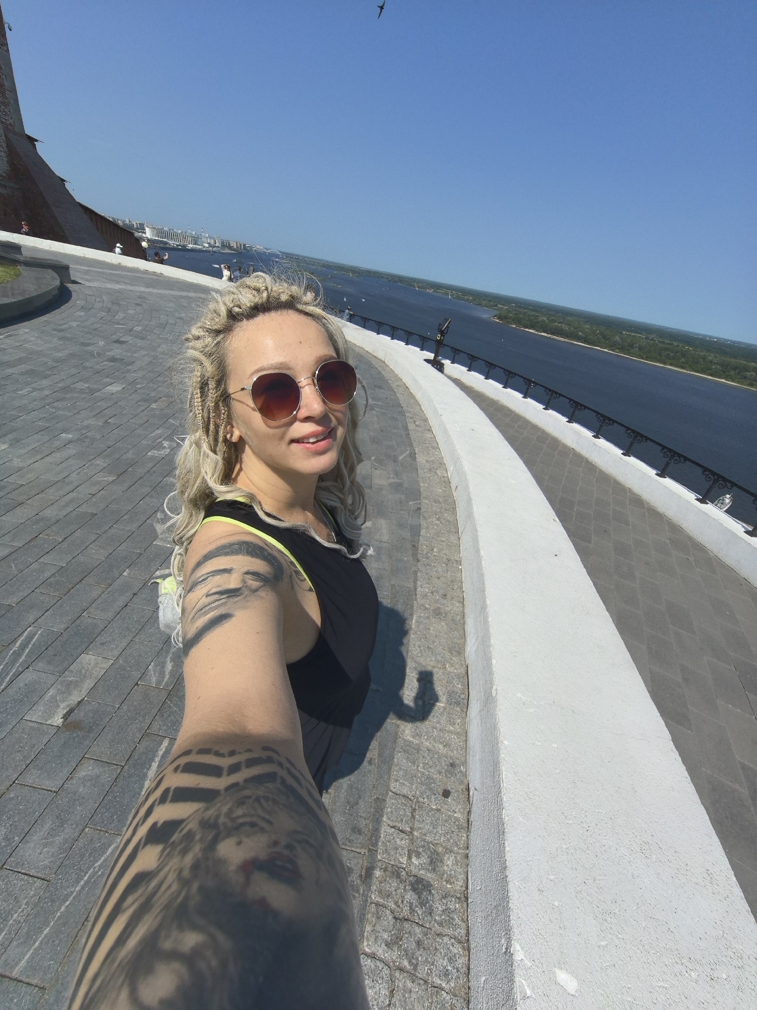 A woman with blonde braided hair and sunglasses takes a selfie in front of a wide river in Nizhny Novgorod, Russia. She is wearing a black tank top with a neon green strap and has extensive tattoos on her arm. The background shows a promenade with a white railing, a paved walkway, and a distant cityscape. The sky is clear and blue.