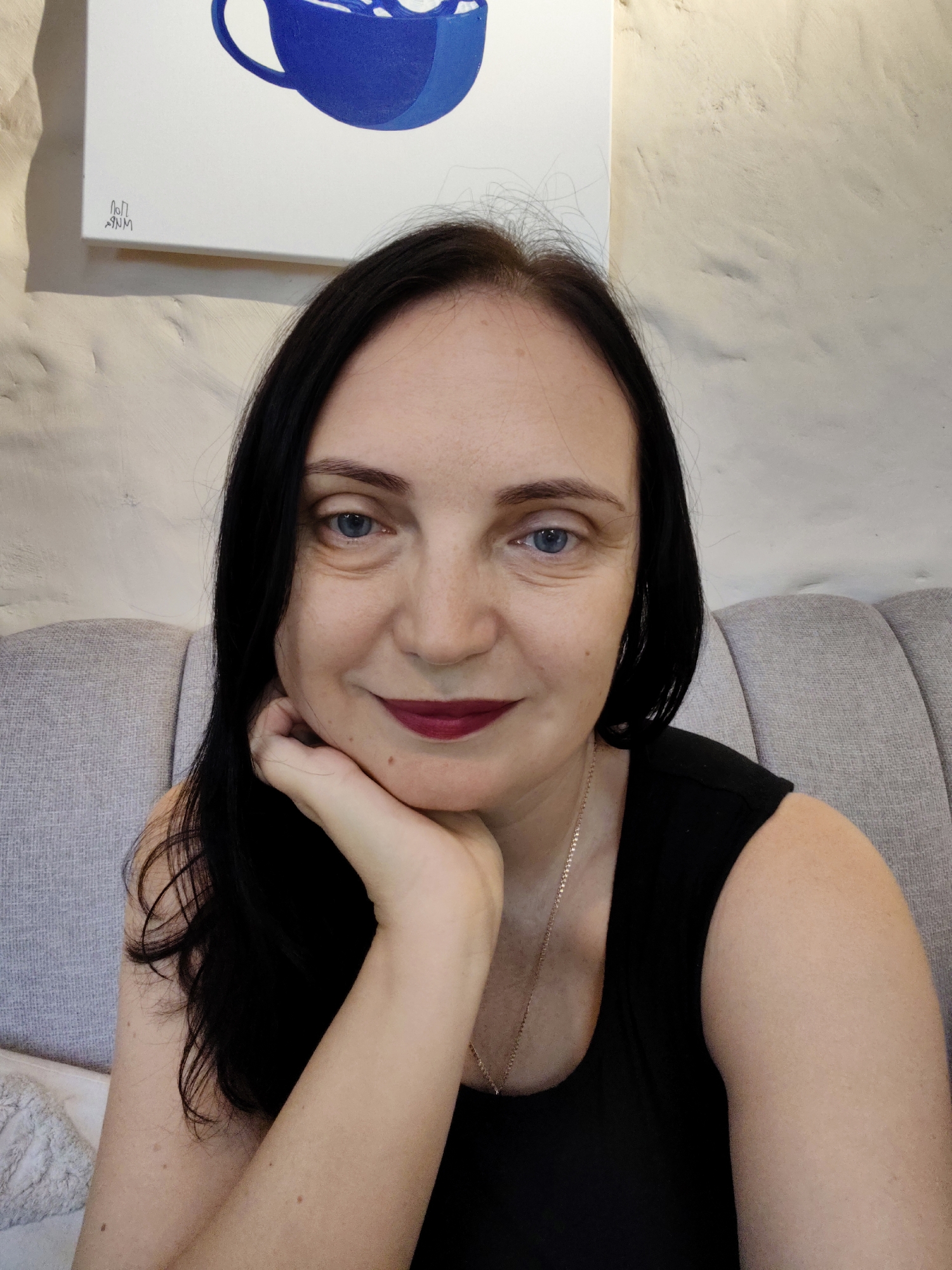 A medium shot shows a Caucasian woman with black hair and blue eyes, wearing a black top and dark lipstick. She is seated on a gray couch, leaning her chin on her hand. A painting of a blue cup with white foam hangs on a textured, light-colored wall behind her. The image does not provide any information about the location within the city of Sochi, Russia.