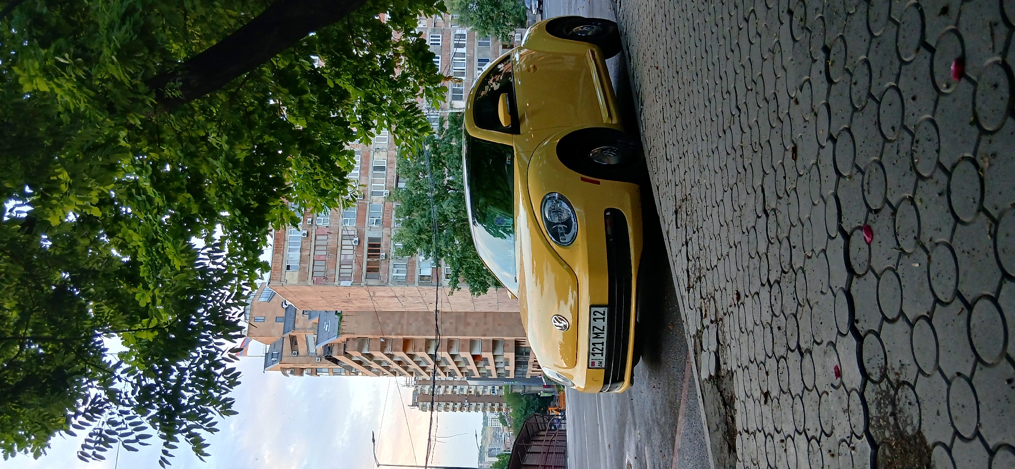 A yellow Volkswagen Beetle is parked on a sidewalk paved with hexagonal concrete tiles. The car faces to the right of the frame, with its front bumper prominently displayed. A license plate on the front reads "AM 121 MZ 12". Behind the car, several tall, beige apartment buildings with numerous windows and balconies dominate the background. A large tree with dense green foliage partially obscures the upper portion of the buildings and the sky. The street in front of the sidewalk appears wet. The scene is set in a city environment, with the architecture suggesting a location in Yerevan, Armenia. The sky is overcast with some visible clouds.