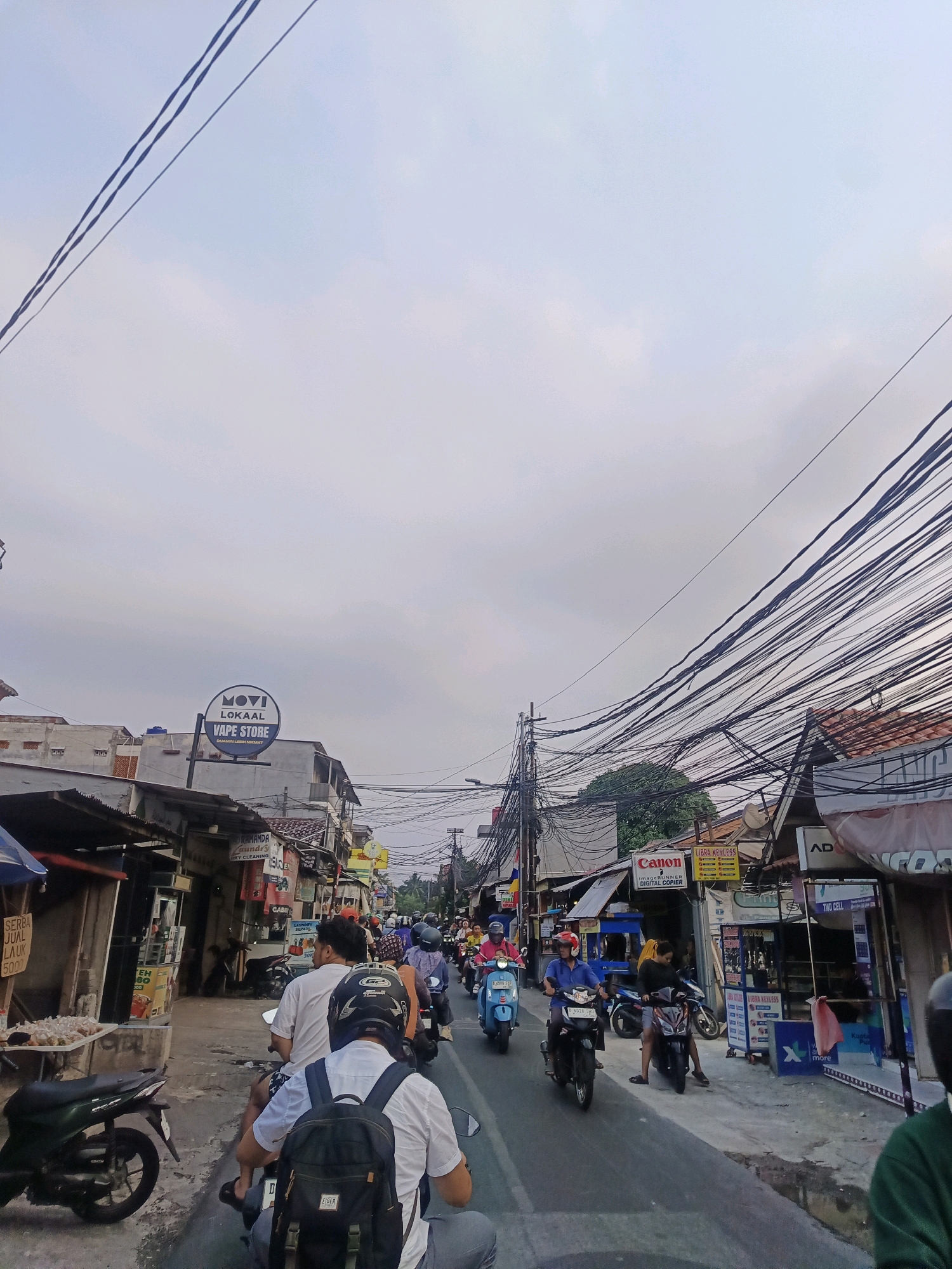 The image depicts a street scene in South Jakarta, Indonesia, likely during daylight with overcast skies. The street is narrow and lined with shops on both sides, with a significant amount of overhead wiring creating a dense network across the sky. A stream of motorcycles and their riders are navigating the road, suggesting moderate traffic.
On the left side of the street, a shop with a corrugated metal awning displays goods on a table, including packaged items labeled "SERBA JUAL LAUK". Further down, a shop sign reads "MOVI LOKAA! VAPE STORE".
On the right side of the street, various businesses are visible, including a "Canon IMAGERUNNER DIGITAL COPIER" sign and another advertising "LIBRA KEYLESS". Other storefronts are present with signage that is partially obscured or difficult to read.
The people in the image are primarily riding motorcycles, many wearing helmets. Some are facing forward, while others are looking around. The overall atmosphere appears to be a busy urban environment typical of a commercial street in Indonesia.