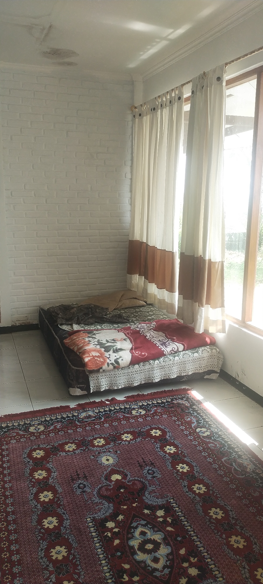 The image depicts a room in a residential building, likely a bedroom, located in Pasirhuni, Indonesia. The room features a low-lying bed with patterned bedding, covered by a floral-patterned blanket. A pillow is visible on the bed. The walls are painted white and appear to be brick construction. A large window with beige and brown curtains is present, allowing natural light to enter the room. A patterned rug is spread across the tiled floor in the foreground. The ceiling shows signs of water damage, with discolored patches. No people are present in the scene.