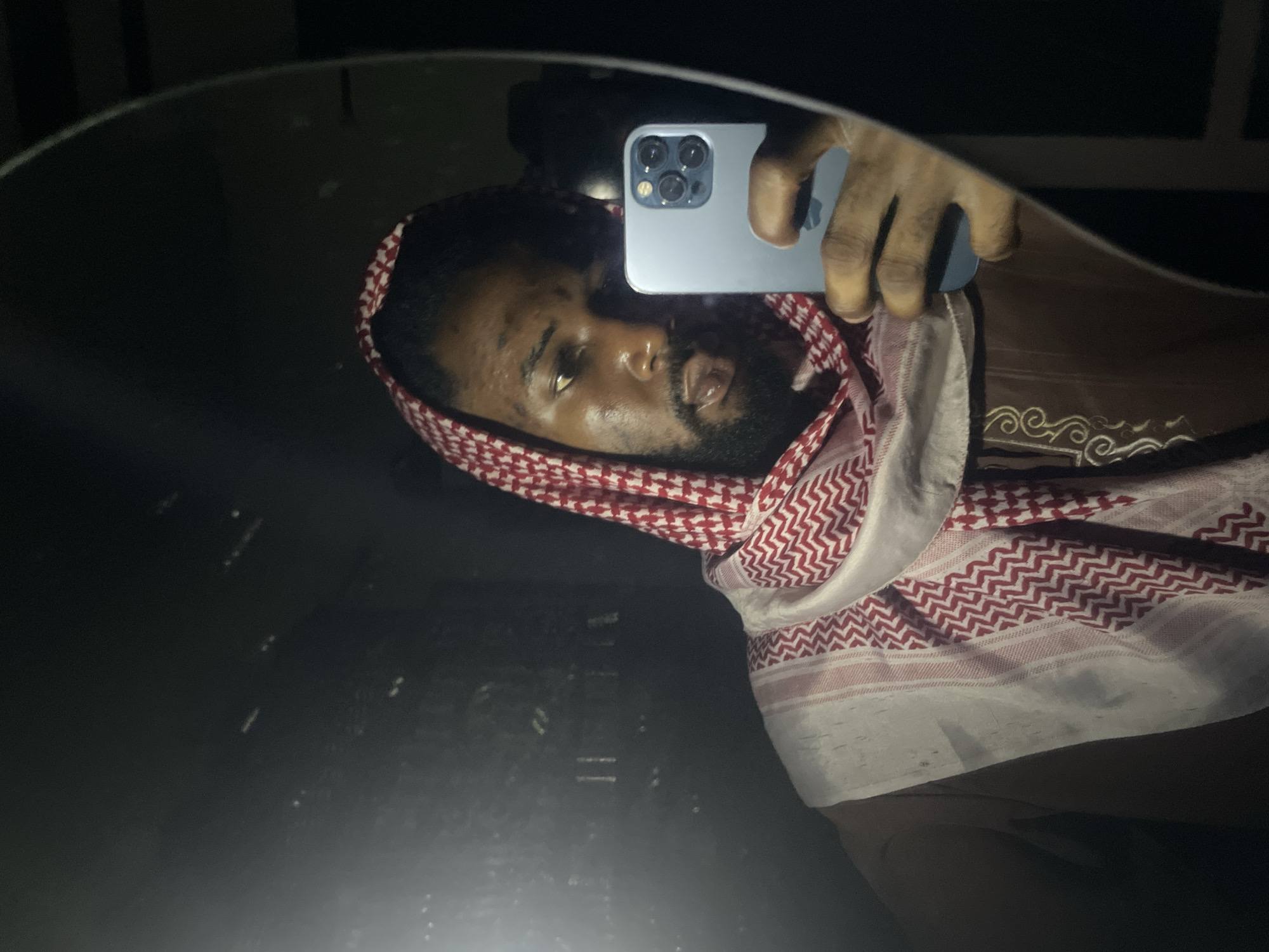 A man with a beard and wearing a red and white checkered headscarf is taking a selfie in a mirror. He is holding a blue iPhone, and the image reflects his face, upper body, and part of his clothing. The background is dark and mostly obscured by the mirror. The location is unknown; there are no identifying features to suggest Lokoja, Nigeria.