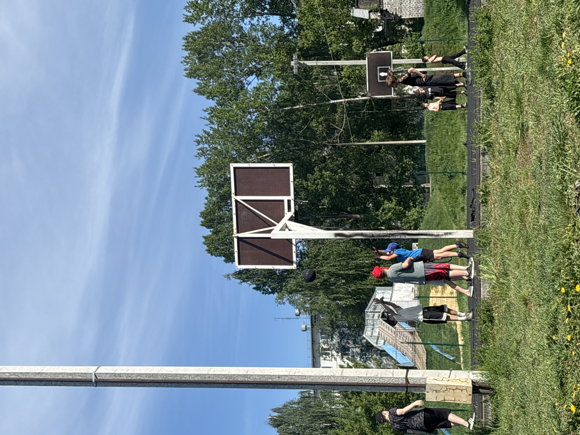 The image shows a basketball court with two basketball hoops. Three people are playing basketball near the main hoop in the foreground. One is about to shoot the ball, one is standing ready to rebound, and one is watching. Two other people are by a second hoop in the background. Tall trees surround the court, and there is a building in the distance. Based on image content, it is possible this image could have been taken in Chaikovsky, Russia, but without further evidence it is hard to confirm.