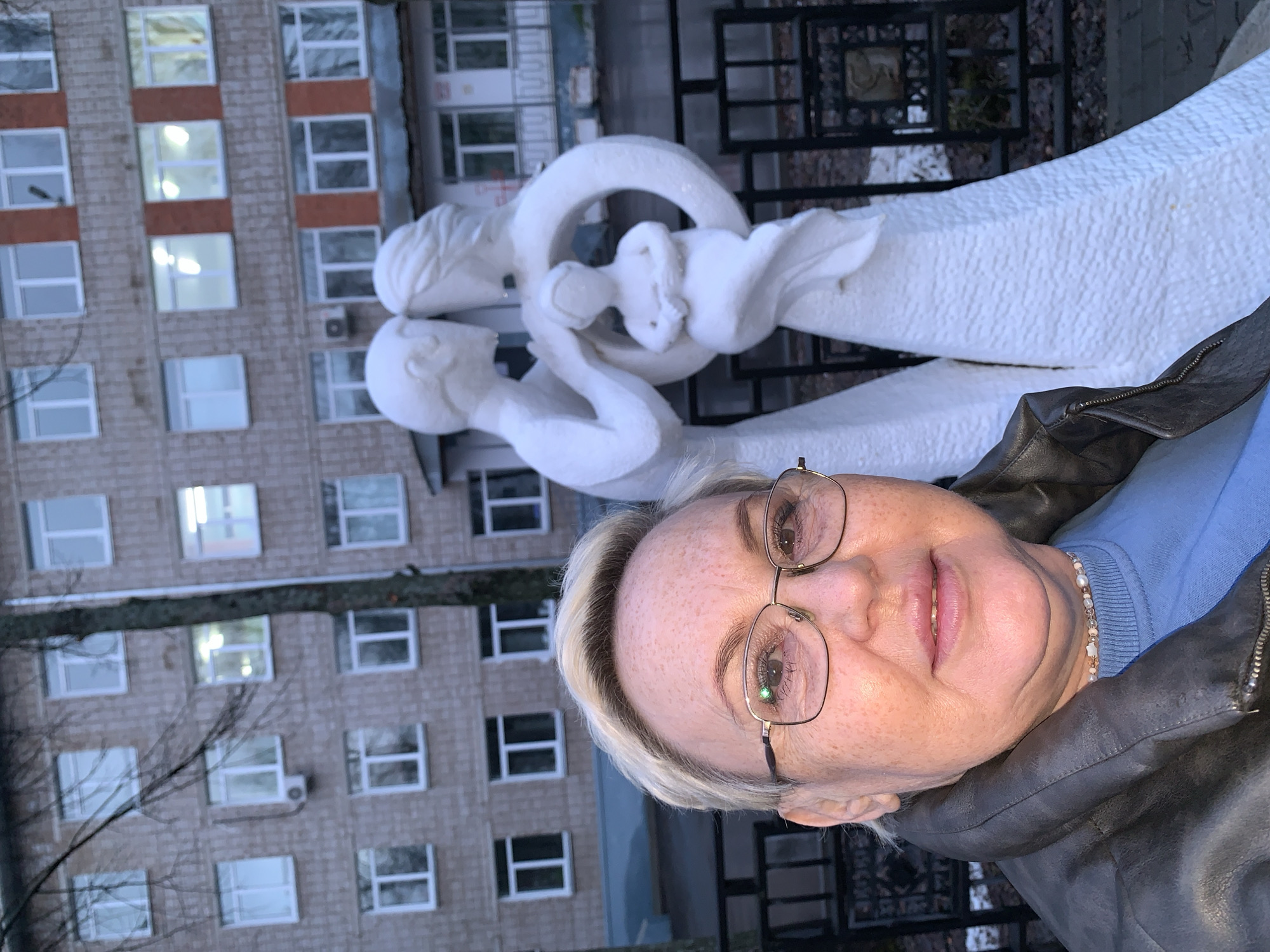 A woman is posing in front of the "Family" sculpture in Chaikovsky, Russia. The sculpture consists of two adults supporting a baby in a circle. A multi-story brick building with evenly spaced windows is in the background. The woman is wearing glasses, a blue sweater, and a leather jacket. The scene appears to be outdoors during daylight hours. A decorative black metal fence is partially visible behind the sculpture. A tree branch is visible in the lower left.