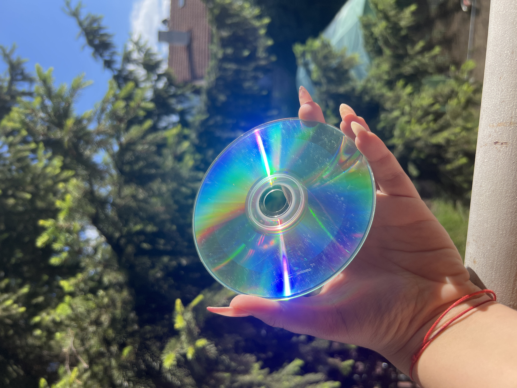 A close-up shot shows a person's hand holding a compact disc (CD) against a backdrop of green foliage, a bright blue sky, and a section of a tiled roof with a chimney, indicative of a residential area. The CD reflects a spectrum of iridescent colors, including blues, greens, and purples, due to sunlight. The person's fingernails are painted a pale pink. A thin red string bracelet is visible on the person's wrist. The background foliage appears to be part of a tree, possibly a spruce, with some areas softly blurred, suggesting it is outdoors. The presence of a tiled roof and chimney strongly suggests an urban or suburban residential setting, possibly in a city like Sofia, Bulgaria, known for its blend of residential areas and green spaces.