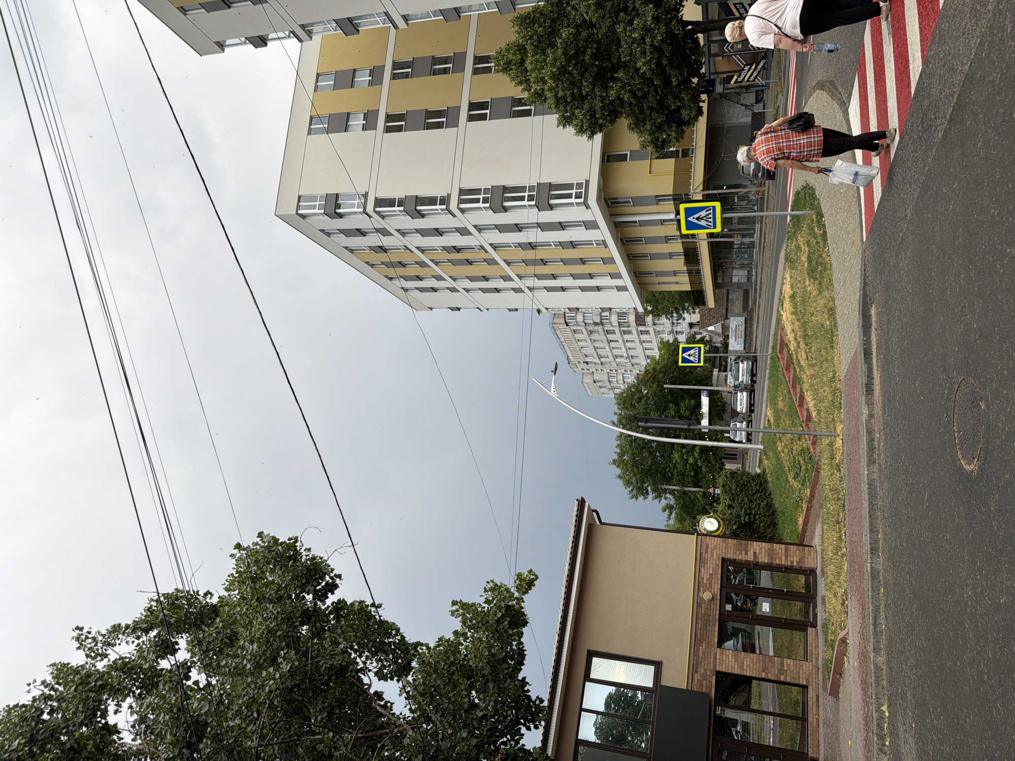 The image depicts a street scene in Strășeni, Moldova. In the foreground, a pedestrian crossing with red and white stripes is visible on an asphalt road. Two women are crossing this intersection. The woman on the left wears a red and white checkered shirt and dark pants, carrying a white shopping bag and a black purse. The woman on the right wears a pink shirt and dark pants, carrying a bottle of water. Behind the pedestrian crossing, a light brown building with a black facade and large windows is located. Further back, a modern apartment building with yellow and white exterior walls and gray window frames stands tall. Adjacent to this building is a dense green tree. In the distance, other residential buildings and more trees are visible under a cloudy sky. Overhead, electrical wires crisscross the sky. Yellow pedestrian crossing signs with blue backgrounds and white pedestrian symbols are placed along the roadside. Several cars are also visible on the street, parked or driving. The overall impression is of an overcast day in an urban environment.