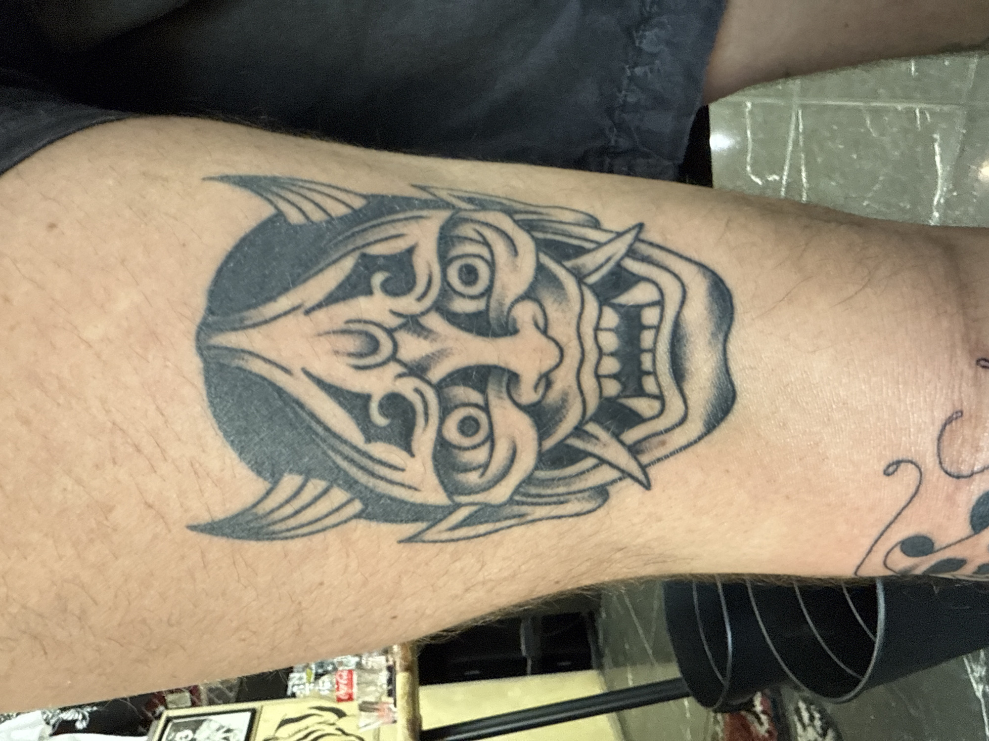 The image shows a person's forearm with a tattoo. The tattoo is a black and white image of what appears to be a Japanese Oni mask. The surrounding area includes skin with hair, and parts of clothing, and furniture. The setting and location of the city of Moscow, Russia cannot be determined from the given image.