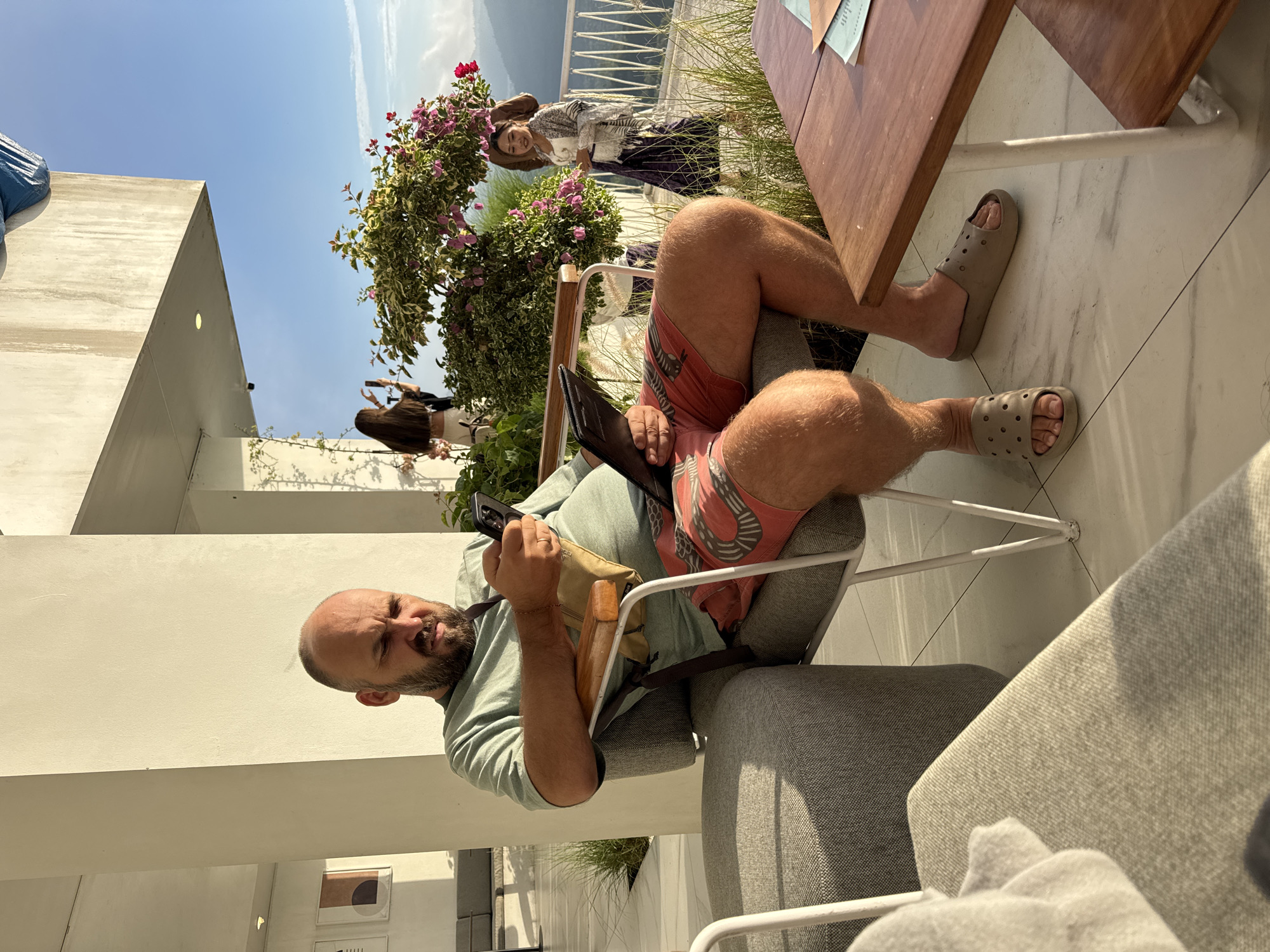 The image depicts a man seated in a grey chair on a patio. He has a bald head, a beard, and is wearing a light green shirt, patterned red shorts, and grey Crocs. He holds a black tablet in one hand and a phone in the other, glancing at the camera. He also wears a tan bag across his body. A brown wooden table is to his right.
In the background are two women, one smiling at the camera, standing near a bush with pink flowers. There is a white wall structure and a view of hills or mountains. The image appears to be taken in Kedisan, Indonesia, based on contextual clues.
