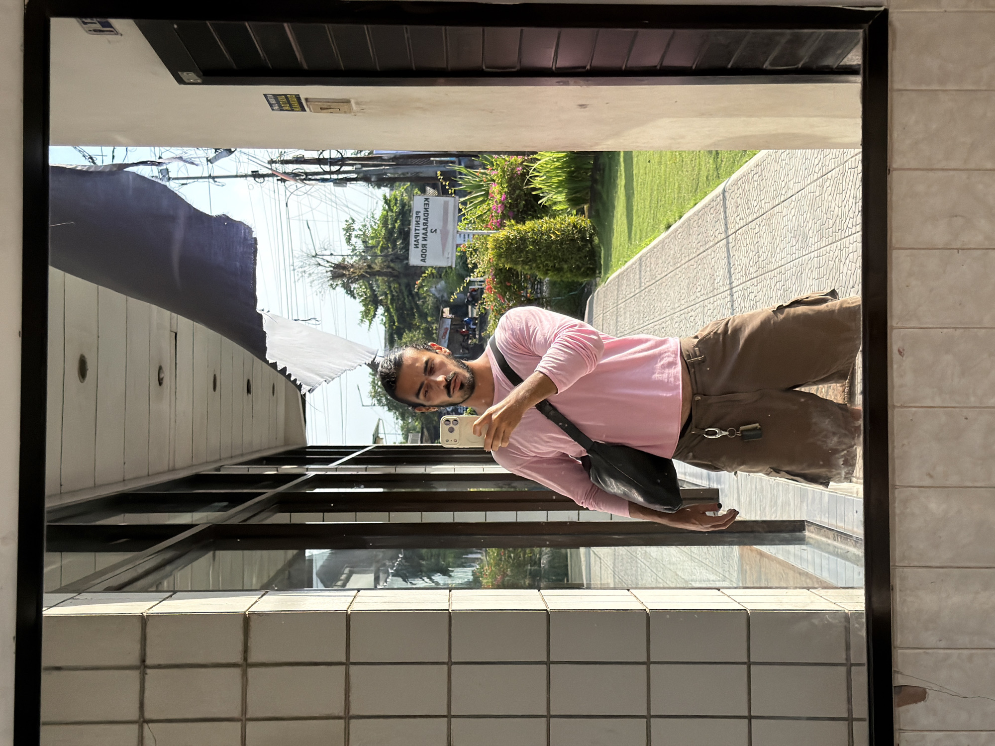 A man is reflected in a large mirror, holding a smartphone and taking a selfie. He is wearing a pink long-sleeved shirt, brown cargo shorts, and a black crossbody bag. A set of keys hangs from his shorts. The mirror is framed by white tiled walls and a dark border. Behind the man, visible through the reflection, is a sidewalk, a grassy area with bushes and flowers, a sign with text in Indonesian, and utility poles with many wires. The scene appears to be outdoors in a built-up area, possibly near businesses or residential buildings. The sign in the background reads "PENILAIAN KENDARAAN PASARAN" which translates to "Vehicle Appraisal Market." This is likely located in Tempurejo, Indonesia, given the Indonesian language on the sign.