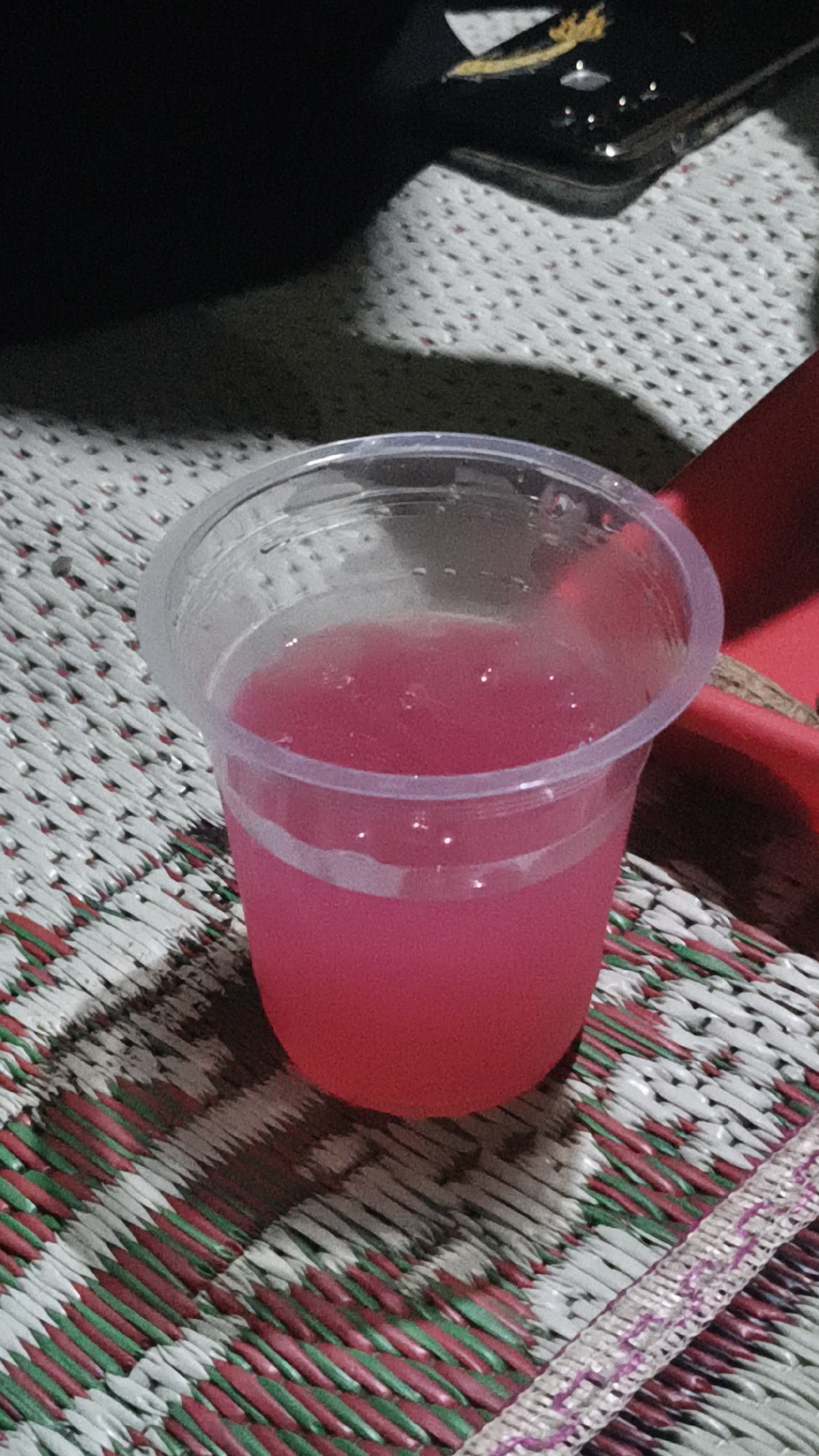A close-up, eye-level shot captures a clear plastic cup filled with a vibrant pink liquid. The cup, slightly wider at the top than the bottom, sits on a woven mat. The mat features a repeating pattern of red and green fibers against a lighter background, with a decorative band running along the lower right edge. Shadows cast by an unseen object create a stark contrast on the mat. In the upper background, a dark, amorphous shape obscures details, with a hint of a metallic object with yellow accents visible. The scene is set in Kerep Kidul, Indonesia.