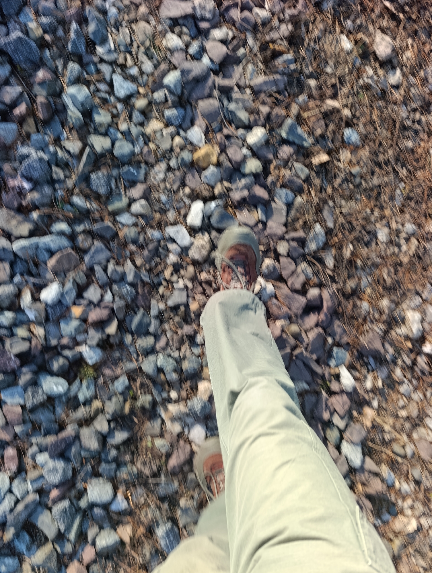 The image shows a first-person view of a person's leg and foot clad in light green pants and a brown shoe stepping on a rocky and dry grassy surface. The ground is composed of a mix of small rocks and dry grasses, with varying colors. The location is potentially outdoors, although the specific setting is unclear without further context. There is no way to know for sure if the location is Krasnoyarsk, Russia.