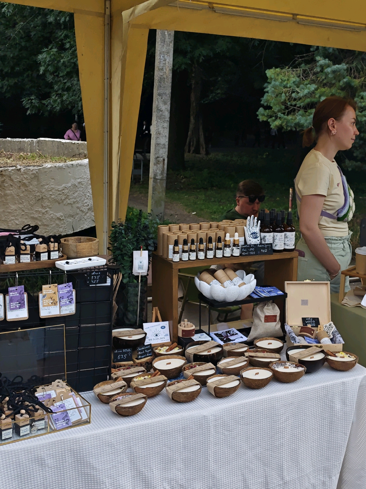 The image depicts an outdoor stall, likely at a market, displaying various handcrafted items. The stall is covered by a yellow awning, and in the background, there are trees and foliage suggesting a park or natural setting. In the foreground, a white tablecloth covers a table laden with products.
Dominating the table are numerous candles presented in coconut shell bowls, some with decorative elements like cinnamon sticks and dried flowers. Several small bottles of essential oils and room sprays are arranged on shelves behind the candles, with labels indicating scents and prices. A wooden box sits on the right side of the table, containing more small items. To the left, a glass display case holds car air fresheners or diffusers.
A woman with her hair tied back in a ponytail, wearing a light-colored t-shirt and pale green shorts, is visible standing behind the stall, looking to the right. Another person, a man wearing sunglasses, is partially visible in the background, appearing to look at the products.
The overall atmosphere is one of a market stall selling artisanal home fragrance products. The provided crops focus on different sections of the stall, highlighting the product displays and the people present. There is no specific visual information to confirm the location in Vinnytsia, Ukraine.