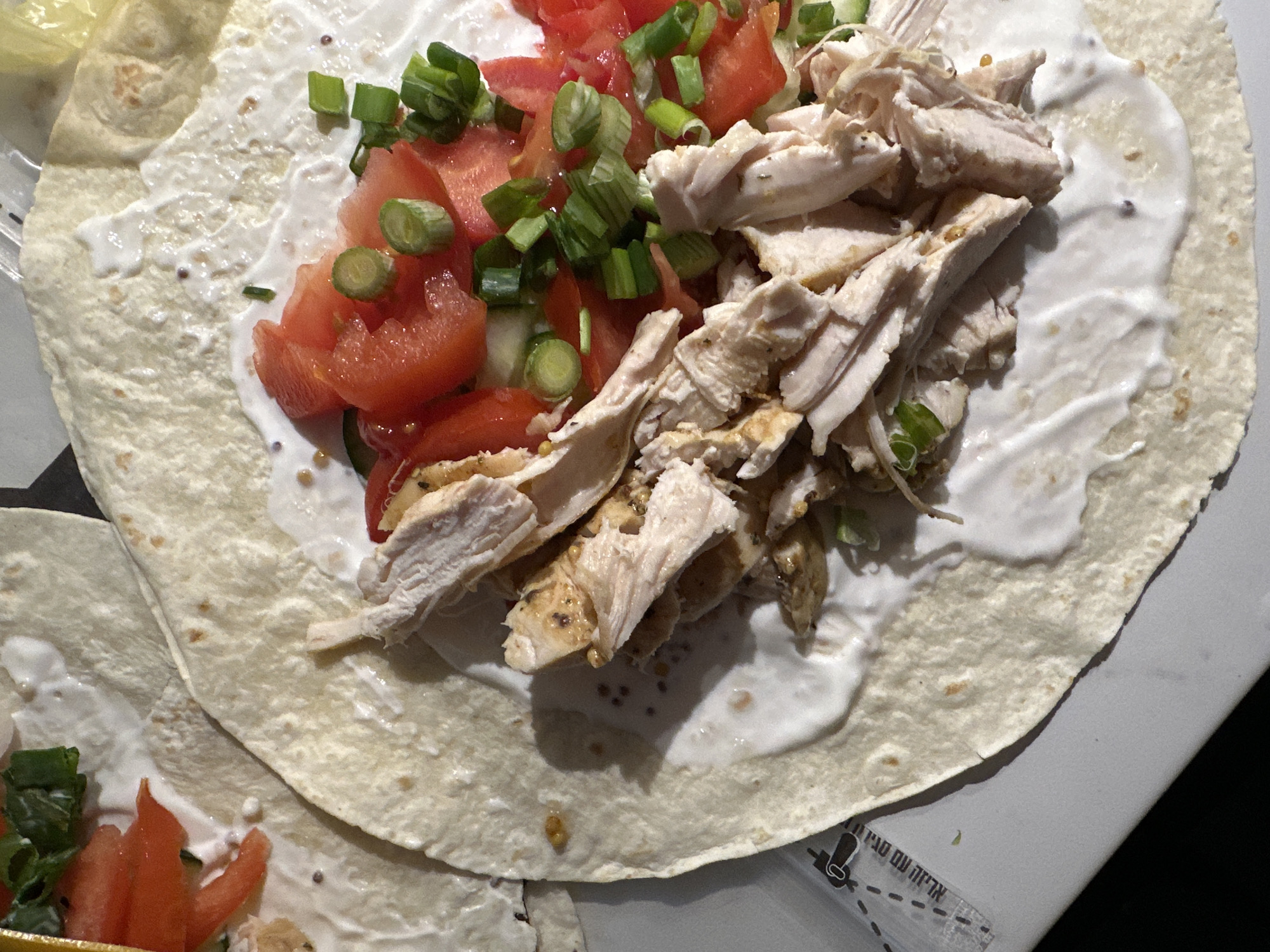 The image shows a partially prepared wrap/burrito. A large tortilla is spread with a white sauce, potentially sour cream or yogurt-based. Diced tomatoes and sliced green onions are layered on one side of the wrap, and shredded chicken is piled on the other. A small portion of another wrap with similar ingredients is visible in the lower left corner of the image.
There are no people visible in the image and there is no information about the location in Budva, Montenegro.