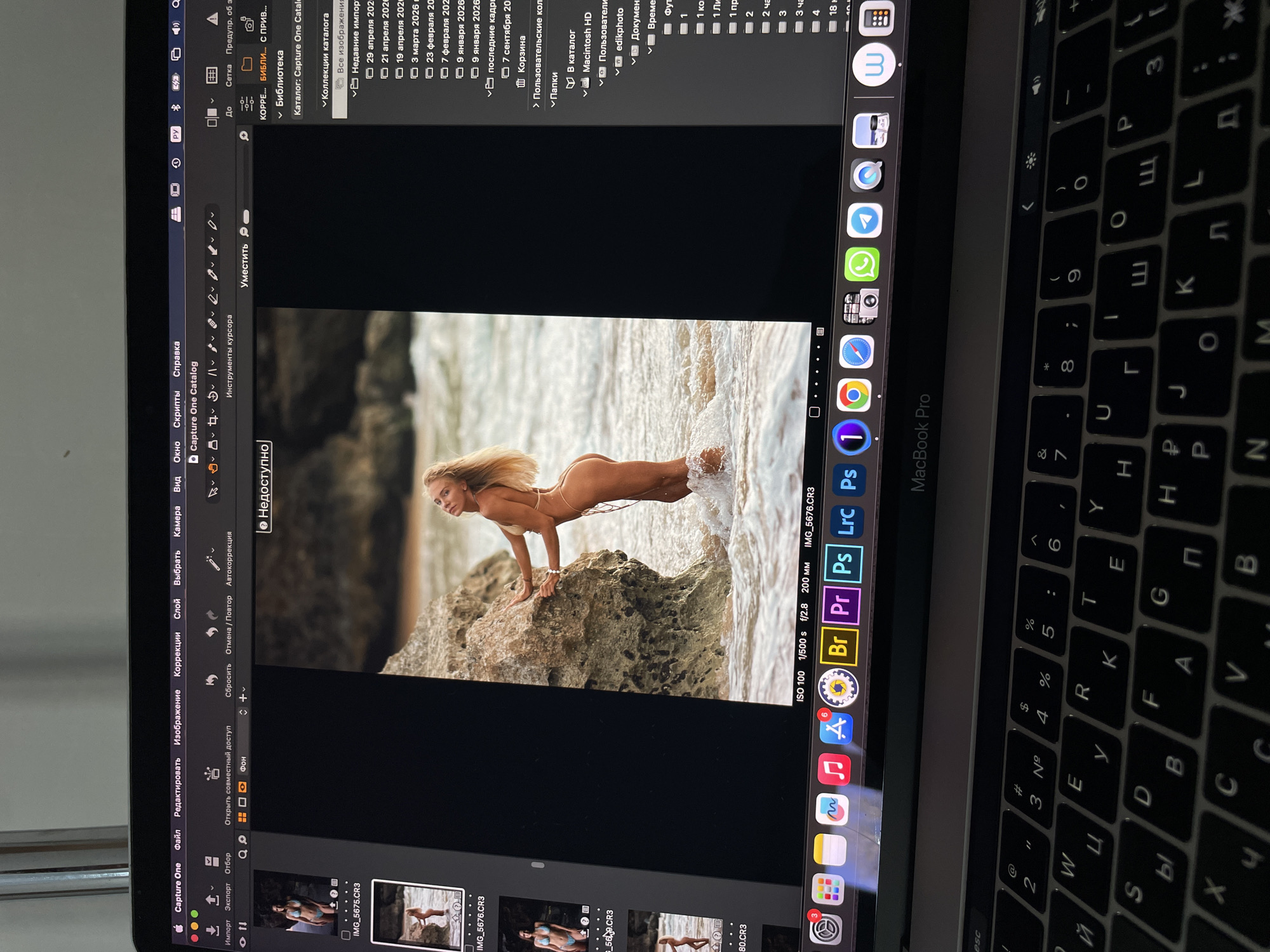 The image is a photo of a MacBook Pro screen displaying the Capture One photo editing software with an image of a woman in a bikini posed on rocks at the edge of the sea. The software interface shows various editing tools and file navigation. The keyboard is visible in the foreground, with Russian/Cyrillic characters alongside Latin ones. The Dock at the bottom of the screen displays various application icons. The image includes some camera settings. There is no indication of the city of Kazan, Russia, in the image.