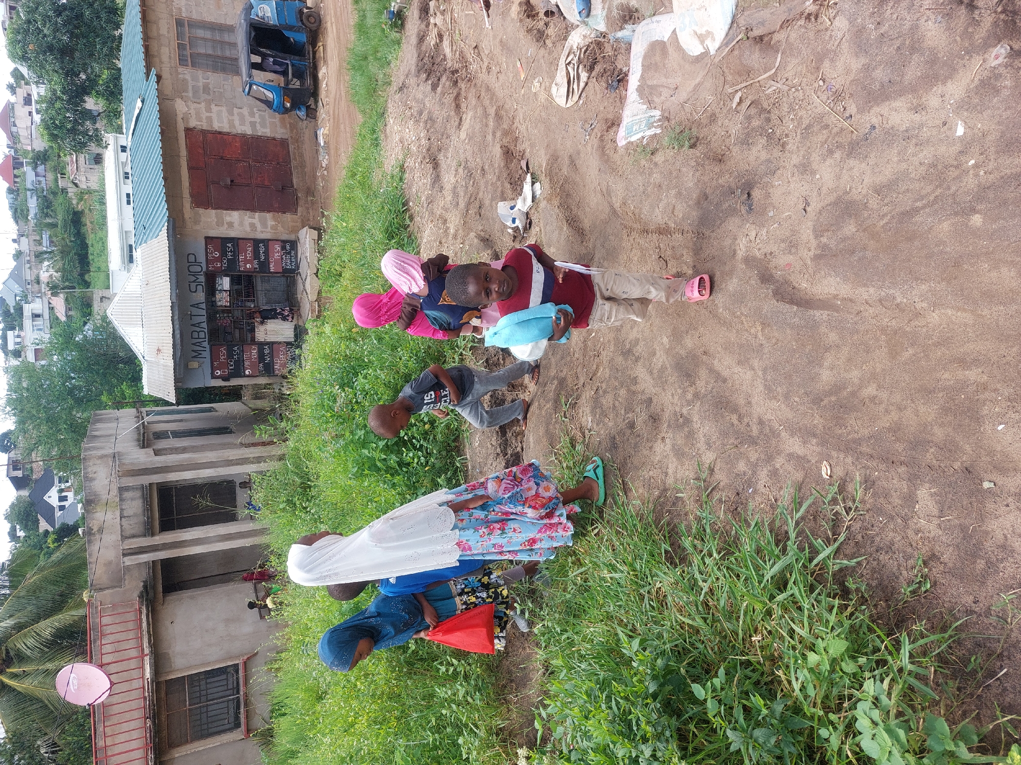 The image shows a group of children standing on a dirt path next to a grassy area, adjacent to a building in Dar es-Salaam, Tanzania.
In the foreground, a group of children, including boys and girls, are the main subjects. Some are wearing traditional head coverings. One boy is holding a blue towel. They appear to be interacting or posing.
Behind the children, there is a dirt path that extends into the background. Scattered on the path are various pieces of trash and debris.
On the left side of the image, there is a building with a faded sign that reads "MABATA SMOP". The building appears to be a small shop or store. Adjacent to this building is another structure, possibly a residential building, with a pink satellite dish attached to it. A three-wheeled vehicle is parked near the store.
The environment is a mix of urban and natural elements. There is green grass and vegetation growing alongside the path. In the distance, there are other buildings and trees.