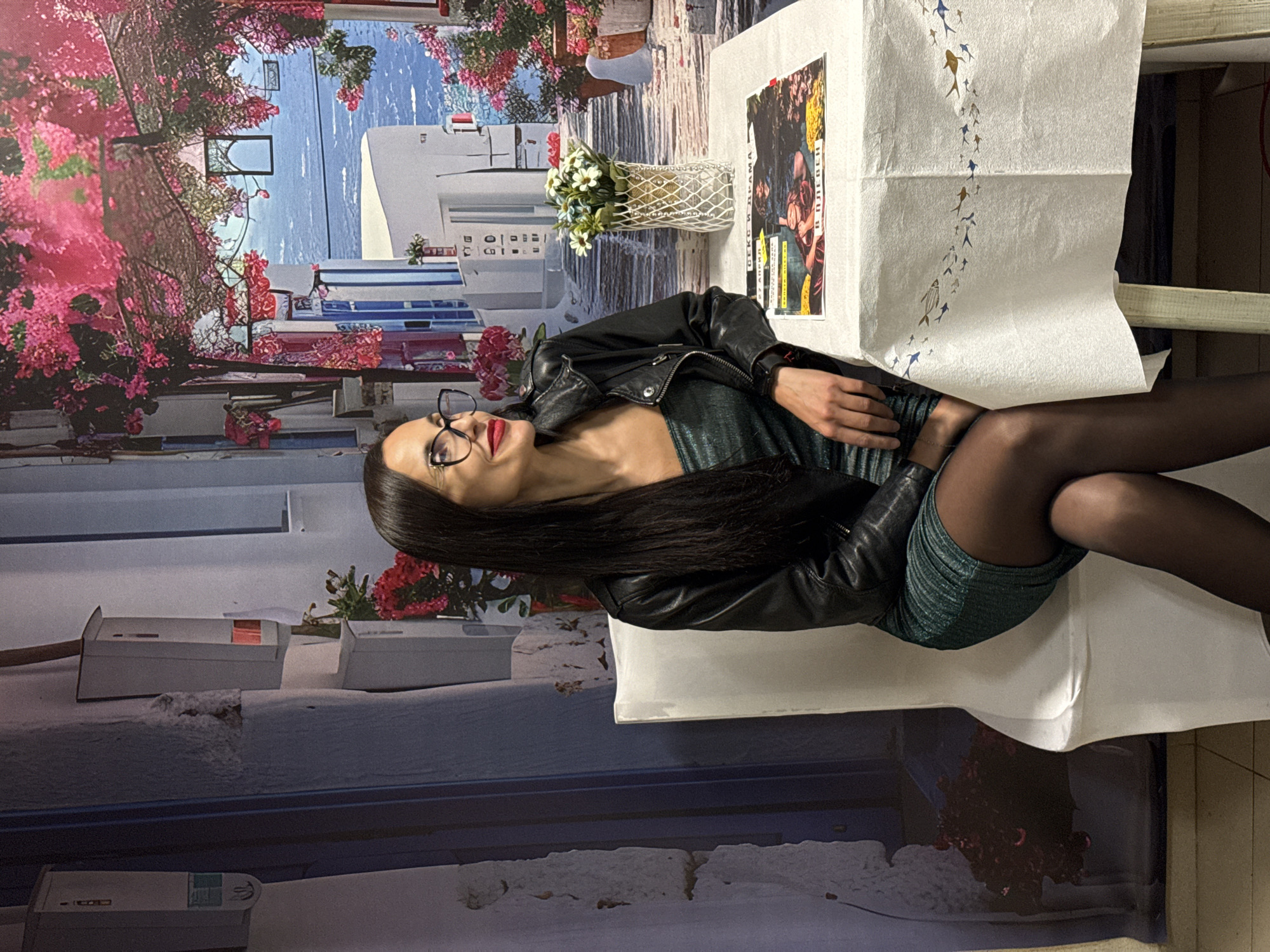 A long-haired woman with glasses and red lipstick sits at a table, wearing a black leather jacket and a green dress. The woman is likely in Pleven, Bulgaria, as indicated by the text on a poster on the table, which reads "В ПЛЕВЕН". The table is covered with a white tablecloth, with a vase of flowers. The background appears to be a wall with painted scenes of buildings, flowers, and a body of water. The woman has black tights.