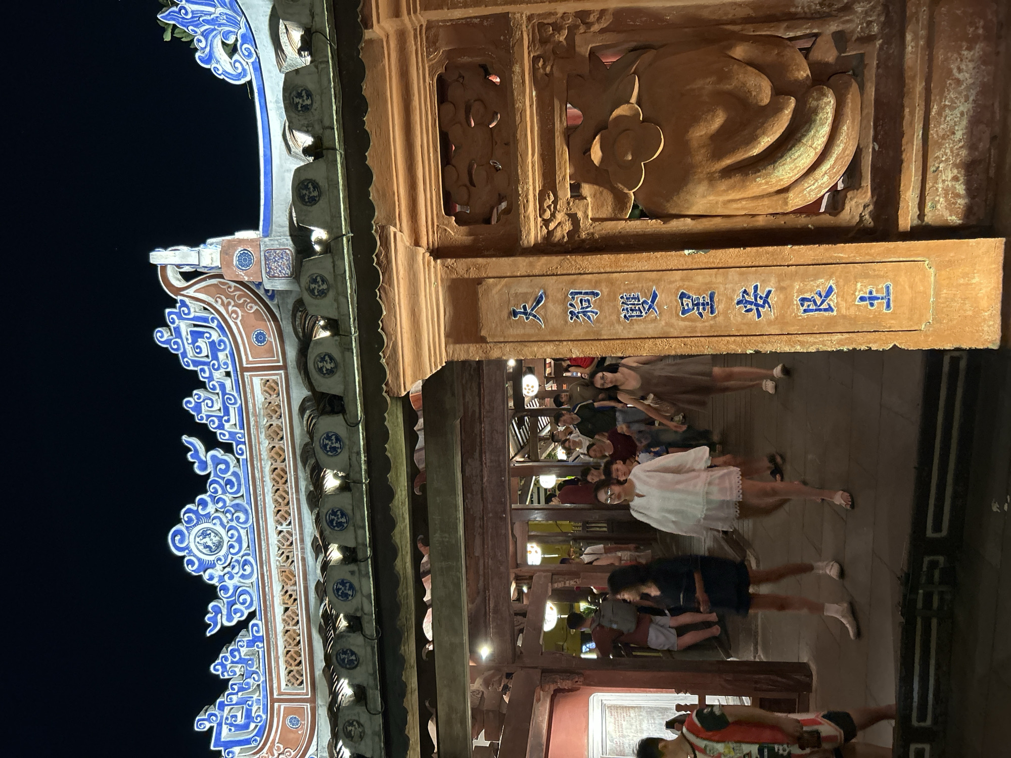 The image shows a nighttime scene in Hội An Ward, Vietnam, with a focus on a traditional architectural structure. The structure features an ornate tiled roof with blue and white decorative elements, and a wooden facade with carved details and vertical script in blue against an orange background. The script appears to be in Chinese characters. Several people are walking through an archway beneath the structure. The individuals are diverse in age and gender, dressed in casual clothing. Some are walking alone, while others appear to be in groups. The lighting suggests an urban environment, possibly a marketplace or a temple complex. The overall atmosphere is one of activity and cultural immersion.