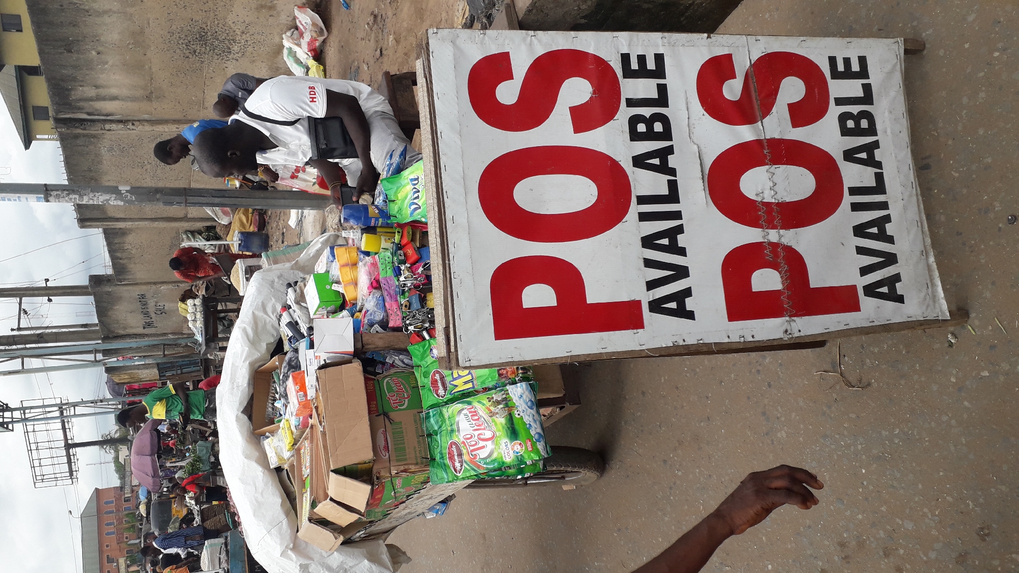 The image depicts a street scene in Onitsha, Nigeria, with a focus on a cart filled with various goods and a prominent sign advertising "POS AVAILABLE." In the foreground, a large white sign with red lettering clearly states "POS AVAILABLE" twice, positioned in front of a wooden cart. The cart is laden with a diverse assortment of items, including boxes, plastic bags, and what appear to be cleaning supplies or detergents, along with smaller packaged goods like keychains or accessories.
Several individuals are present in the background and mid-ground. A man wearing a white t-shirt with the inscription "HDB" is seated on a wooden stool, looking towards the left of the frame. He has a dark bag slung across his chest. Behind him, another man in a blue shirt is partially visible. Further in the background, several other people are engaged in activities typical of a market environment, some standing, some near stalls, and one person appears to be working with goods on a table.
The setting is an outdoor area with a rough, paved surface. A concrete wall runs along the right side of the image, with some graffiti visible, including the phrase "THIS LAND IS NOT FOR SALE". The environment suggests a bustling market or roadside vendor area, with a mixture of goods, people, and infrastructure elements such as poles and overhead wires. The overall impression is one of commerce and everyday life in an urban Nigerian setting.