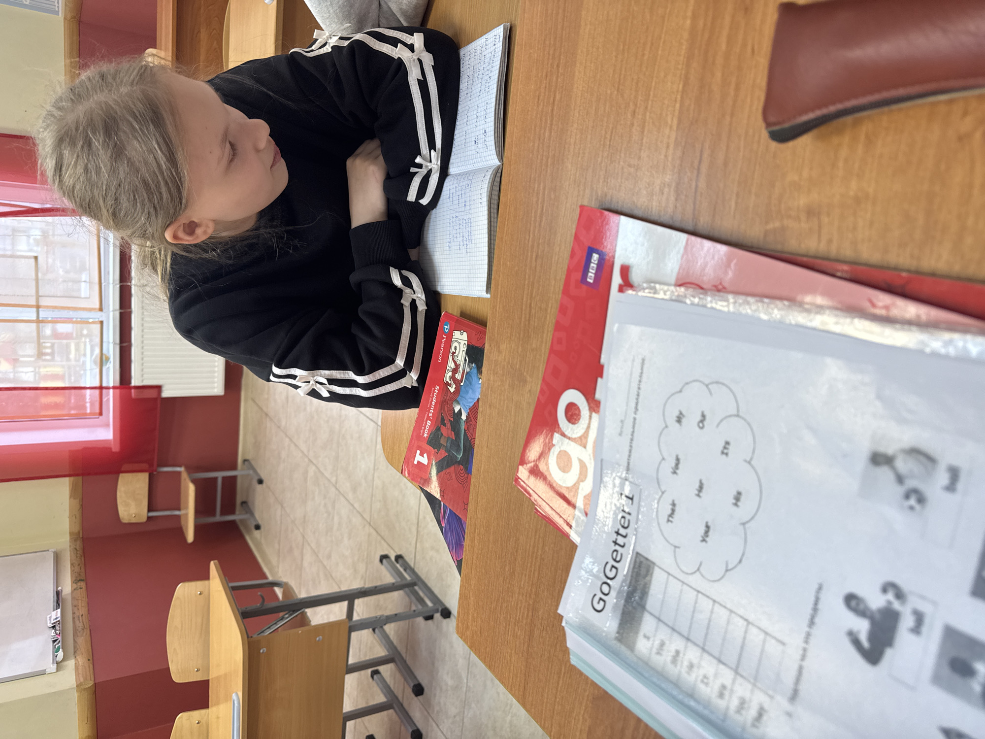 A young girl sits at a school desk in what appears to be a classroom in Perm, Russia. She is wearing a black jacket with white stripes and is resting her head on her crossed arms atop an open notebook filled with handwriting. Several textbooks, including "Go Getter 1," are stacked on the desk. Educational materials with English text are also visible. The classroom features red walls and tile flooring, and additional empty desks are present. A window with a red-tinted pane is partially visible, and a whiteboard is against the wall.