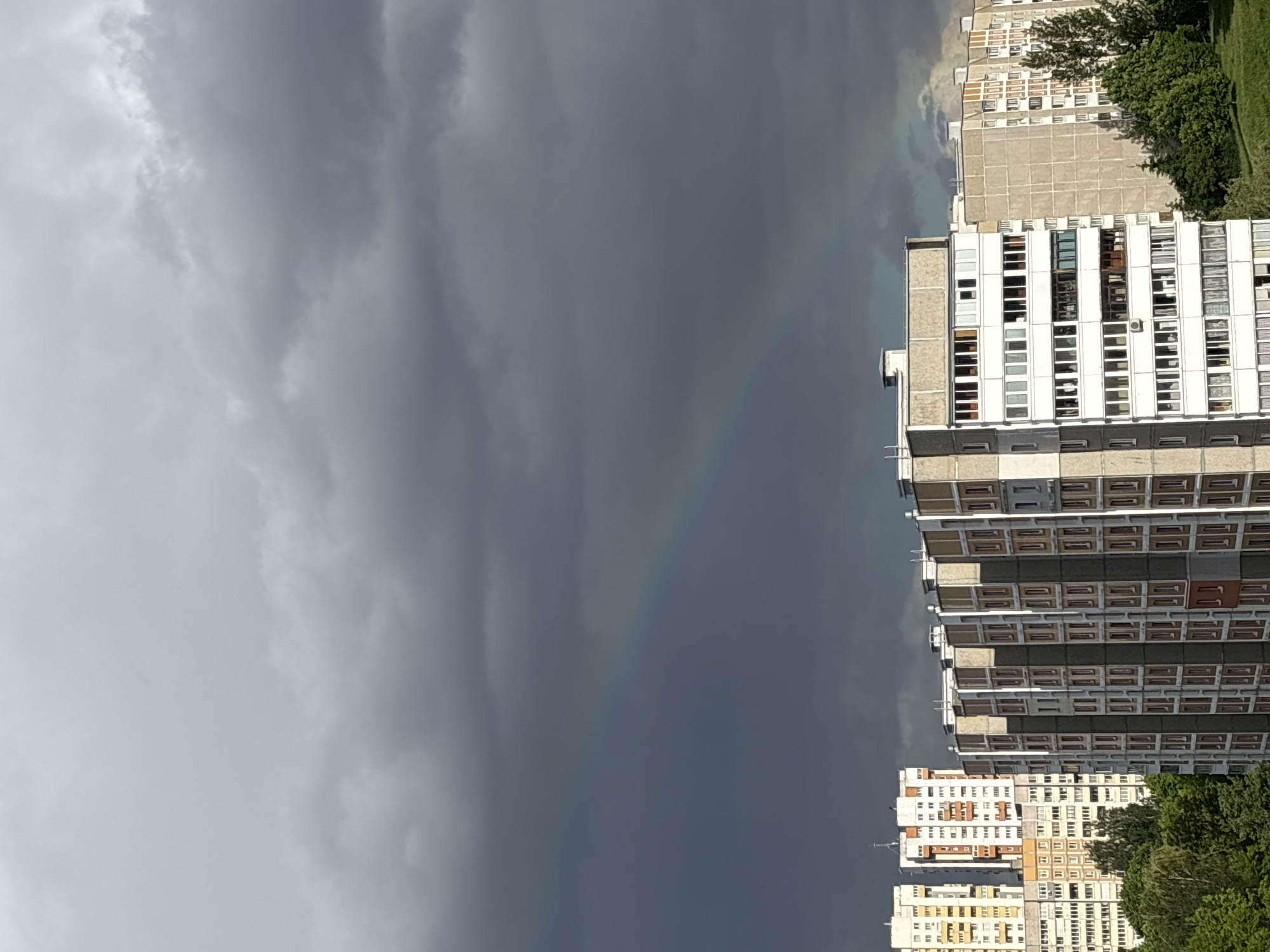 The image shows a cloudy sky over a city. There is a faint rainbow visible against the dark clouds. In the foreground, there are tall buildings with many windows. Trees are also visible next to the buildings. The image is taken in the city of Minsk, Belarus.