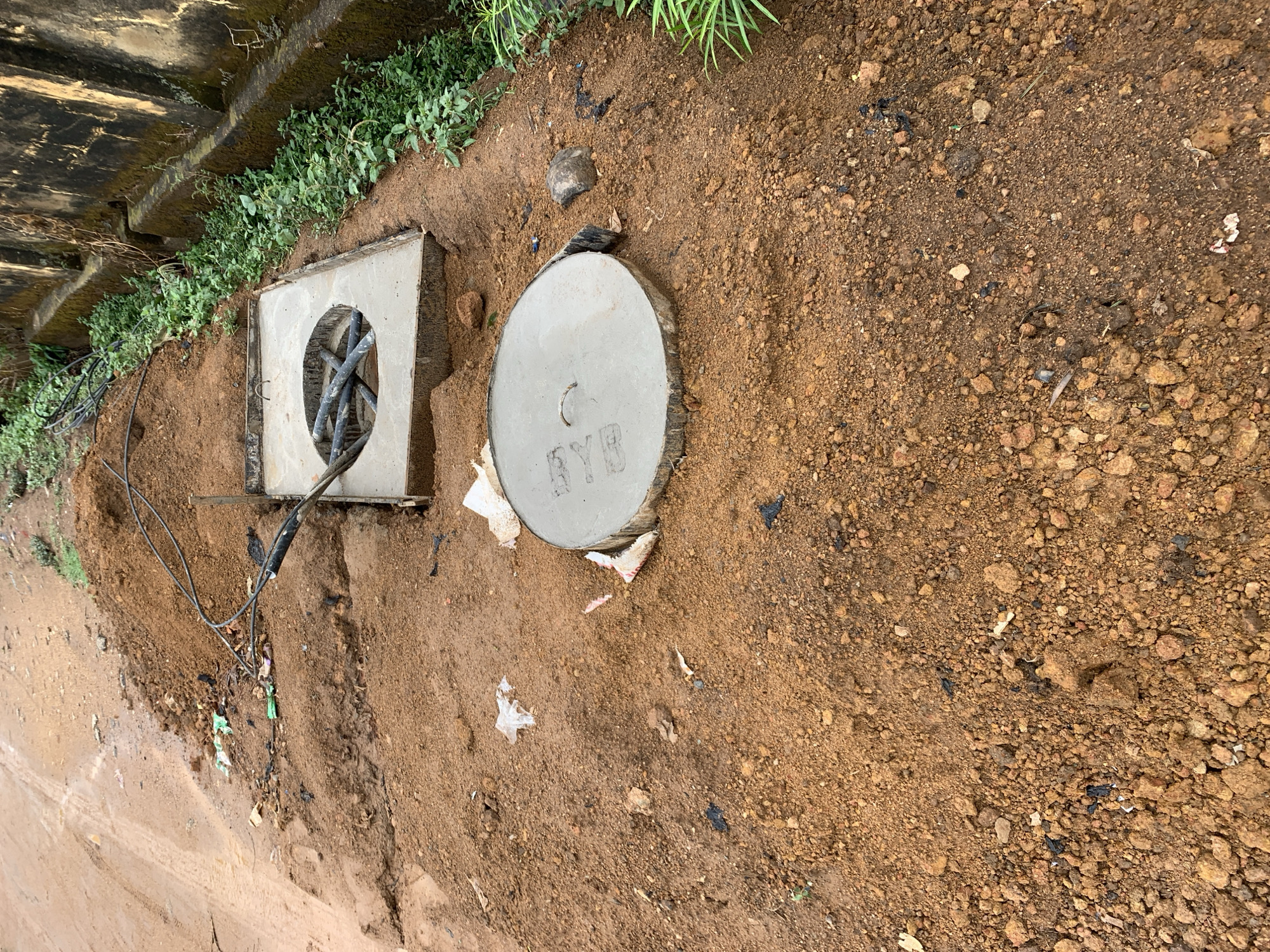 The scene is an outdoor ground-level view next to a structure (possibly a bridge or building) showing utility access points in Rafin Par, Nigeria. The ground is primarily a mix of reddish-brown soil and small rocks. Located at the edge of the structure is a square concrete frame with a circular opening containing several black cables bundled together. Adjacent to it is a round concrete manhole cover with the inscription "RYB" and a slot-shaped indentation. There is vegetation growing along the structure’s edge. Debris, including what appears to be plastic scraps, are scattered on the ground. The overall impression is one of infrastructure and utility access in a semi-developed area.