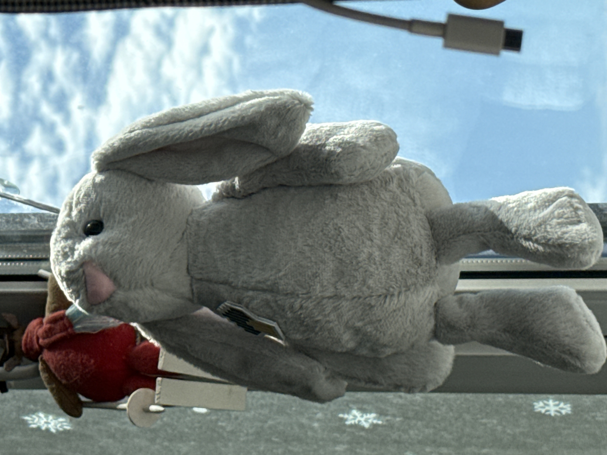 A plush grey bunny with long ears is hanging in front of a window. The bunny has pink accents on its nose and ears. Behind the bunny, a red stuffed toy with brown antlers is visible, partially obscured. Snowflakes are visible on the window. The background is a bright blue sky with white clouds, suggesting an outdoor view. A white charging cable with a USB connector is also visible on the right side of the frame, hanging near the window. The setting appears to be indoors, looking out a window, possibly in a residential or office space. The image does not provide any information to determine a specific location in the city of Krasnoyarsk, Russia.