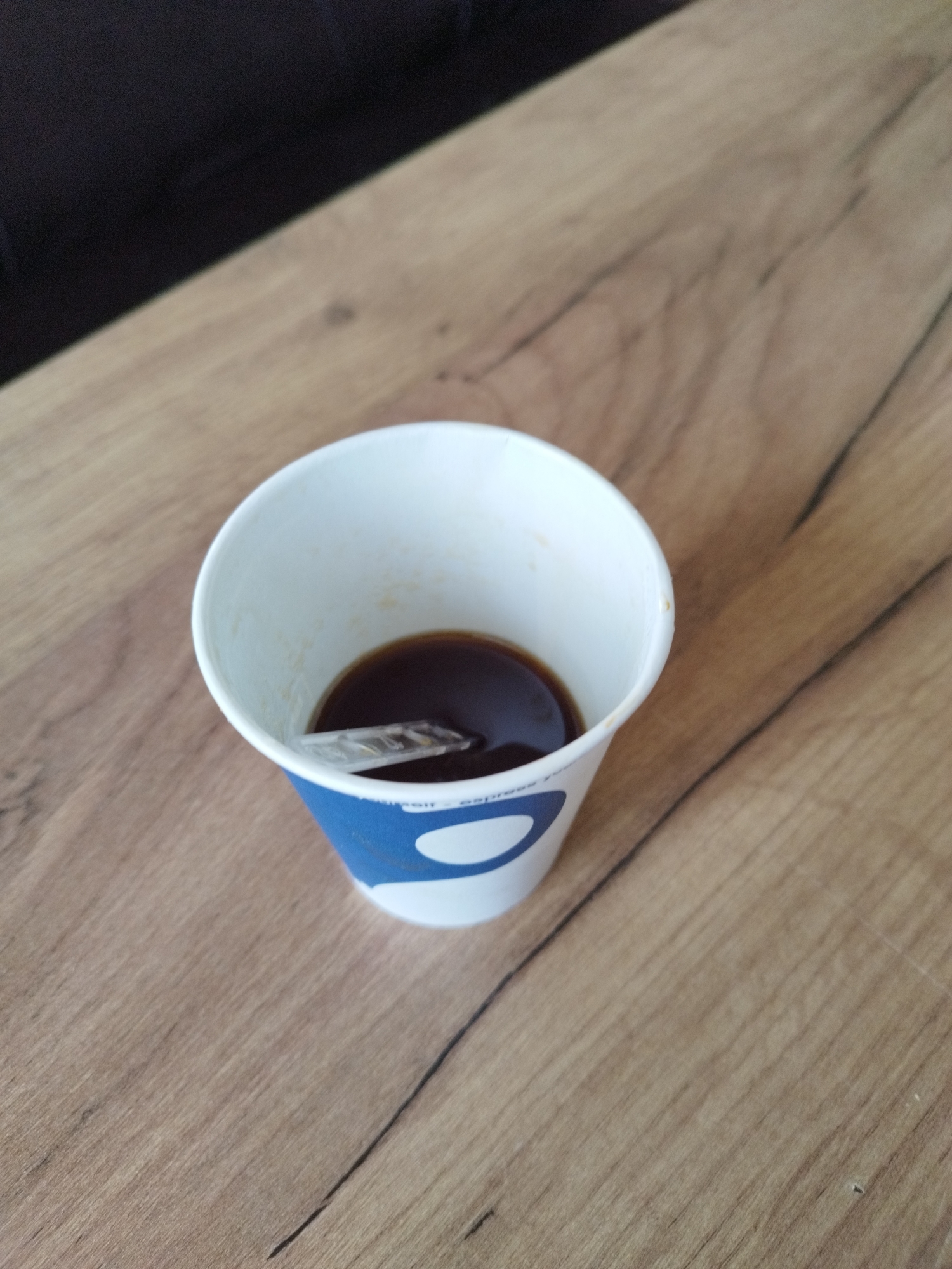 A high-angle shot depicts a white paper cup, blue at the base, sitting on a light brown wooden surface. Inside the cup, a small amount of dark liquid, possibly coffee, is visible, with a plastic spoon resting within. The background consists of the same wooden surface and a dark, blurry object. The location appears to be indoors, possibly a cafe or home setting.