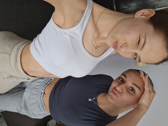 Two young adult females are depicted indoors in what appears to be a residential or private setting within Sofia, Bulgaria. The lighting is bright and even, suggesting an overhead light source or indirect natural light.
The female on the left is positioned slightly in front, facing forward, and is shown from the chest up, though her lower torso and legs are also partially visible. She has light skin, brown hair, and appears to be winking with her right eye while puckering her lips. She wears a light grey ribbed crop top, two gold chain necklaces, and has earrings in her left ear, including a hoop and a stud. Her lower body is clad in beige ribbed sweatpants.
The female on the right is partially visible behind the first, also facing forward and looking towards the camera. She has darker skin, dark hair, and is puckering her lips while holding her left hand against the top of her head. She wears a dark grey t-shirt and light grey sweatpants, the waistband of which displays the text "FAMILY DISAPPOINT." She has a silver chain necklace with a pendant.
The background consists of a plain, light-colored wall on the upper portion, with a visible light switch or electrical outlet on the wall between the subjects' heads. The lower portion of the background, beneath the subjects, shows a dark floor. No specific identifying landmarks of Sofia, Bulgaria are visible within the image, but the location is specified as within the city of Sofia, Bulgaria.