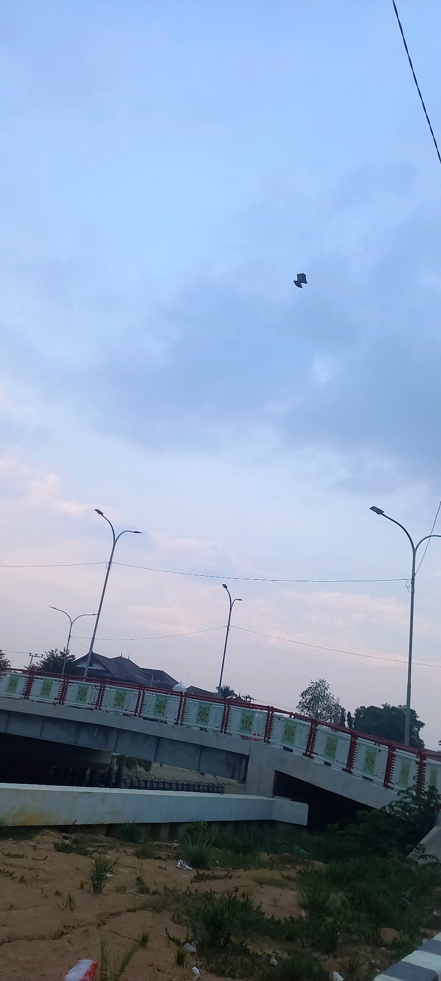 In Melayu, Indonesia, a bridge under construction or recently completed is visible under a hazy, overcast sky. The bridge features a red and white railing with a decorative pattern. Lampposts are positioned along the bridge. In the foreground, sandy ground is scattered with some sparse vegetation and debris. In the upper portion of the image, a kite is flying against the sky. The overall scene suggests an outdoor environment, likely in a developing or suburban area.