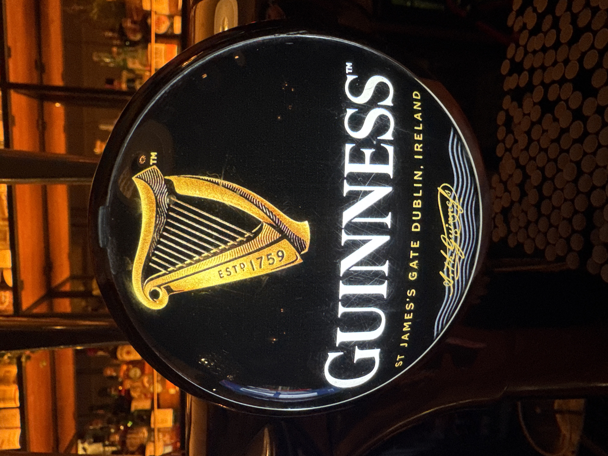 A close-up shows a Guinness beer tap handle. The tap handle is black and circular, with the Guinness logo prominently displayed. The logo includes a gold harp on the left and the name "GUINNESS" in large, white, sans-serif letters on the right. Below "GUINNESS," the text "ST. JAMES'S GATE DUBLIN, IRELAND" is printed. There is also a gold signature underneath this text. The background behind the tap handle is blurry, but it appears to be a bar setting. In the background on the left, a shelf with bottles is visible. On the right, a textured wall or surface with dark dots can be seen. The overall lighting is warm and slightly dim, suggesting an indoor environment.
