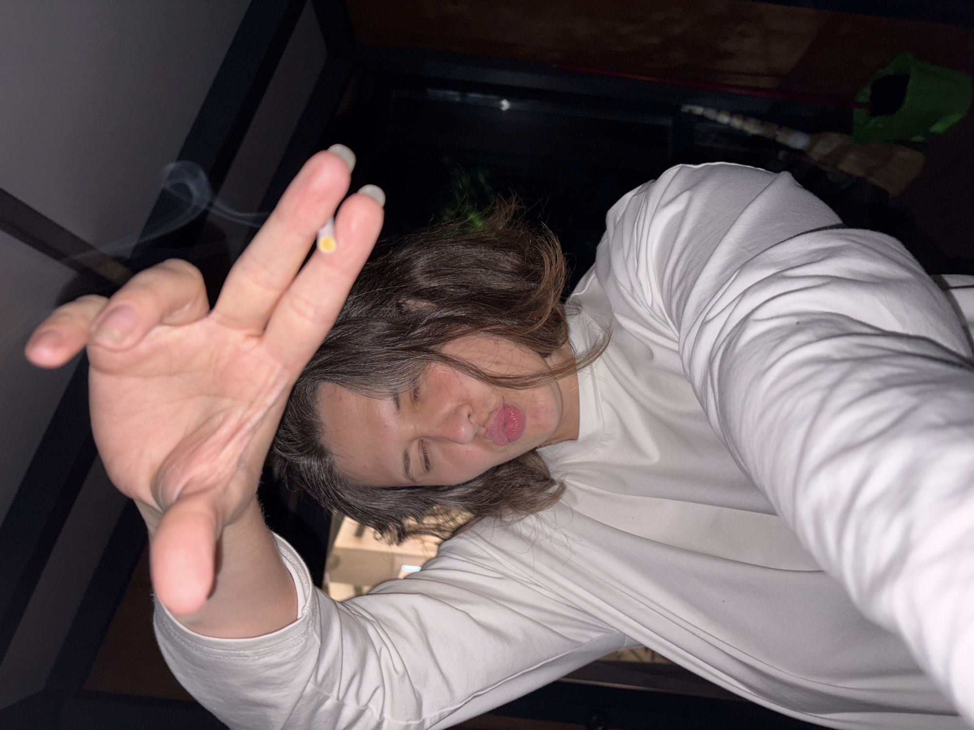 The image shows a close-up, selfie-style shot of a person holding a lit cigarette in their right hand, with smoke rising. The person is wearing a white long-sleeved shirt and has their eyes closed, puckering their lips in a kiss-like gesture. Their left arm is raised and outstretched towards the camera, with the hand open and fingers splayed. The background is dimly lit and appears to be indoors, possibly a porch or patio area, with dark horizontal beams visible overhead. A broom and a green trash bin are partially visible in the lower right corner. The scene is not in Костоваты, Russia.
