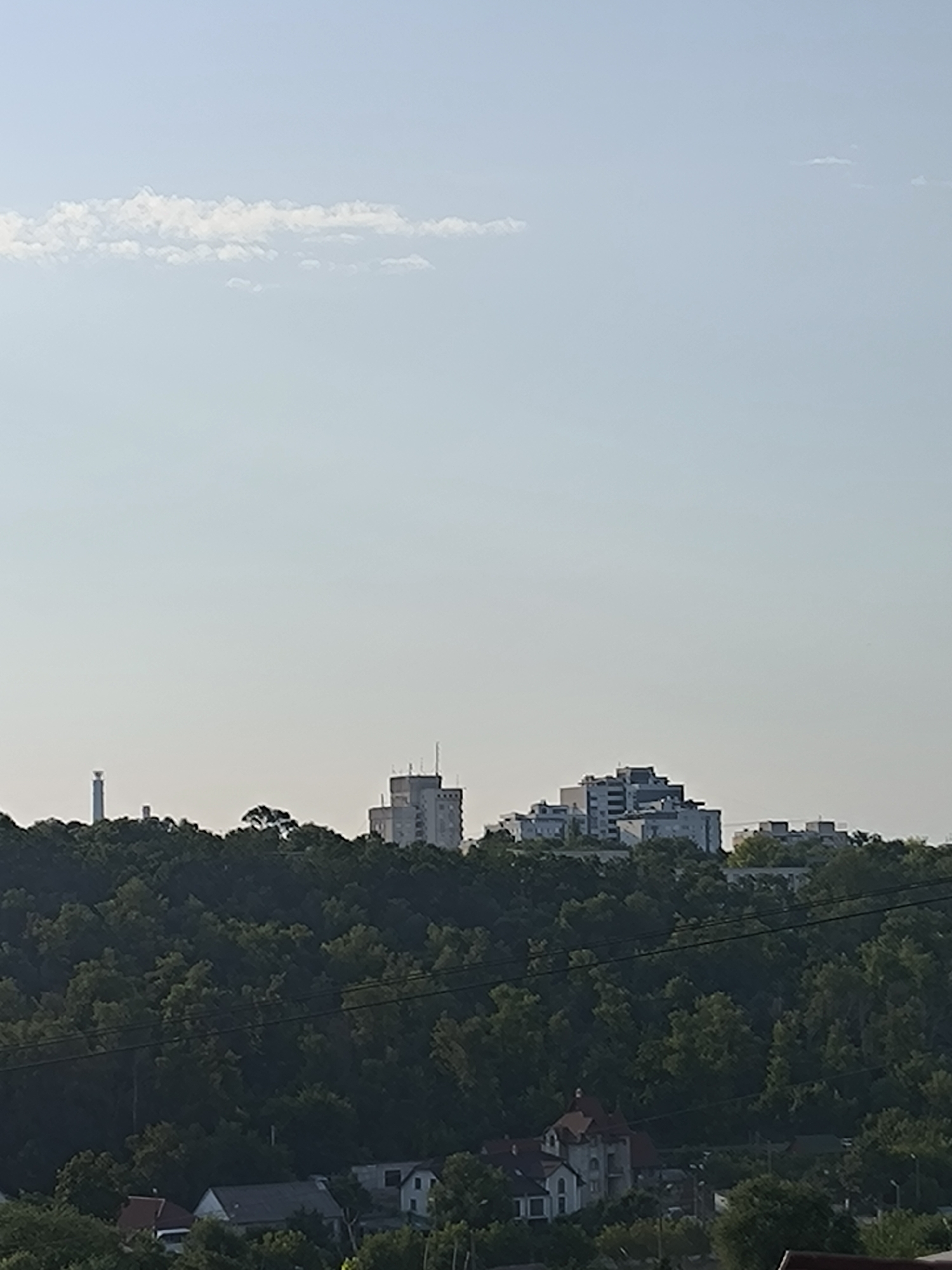This image depicts a cityscape, likely taken from an elevated position overlooking a valley or hill. In the foreground, a dense green forest covers a hillside, with the tops of trees and some residential buildings visible. Further in the distance, atop the hill, several modern, multi-story buildings are visible, including one with a distinct stepped architectural design. To the far left, a tall, slender tower, possibly a lighthouse or radio tower, stands against the pale sky. The sky itself is a light blue with a thin, wispy cloud stretching horizontally across the upper portion of the frame. The overall impression is one of urban development nestled within a natural landscape. The presence of the specific architectural style of the buildings, particularly the stepped design, combined with the terrain and the tower's appearance, strongly suggests this scene is the Râșcani district of Chișinău, Moldova, known for its hilly landscape and distinctive apartment blocks.