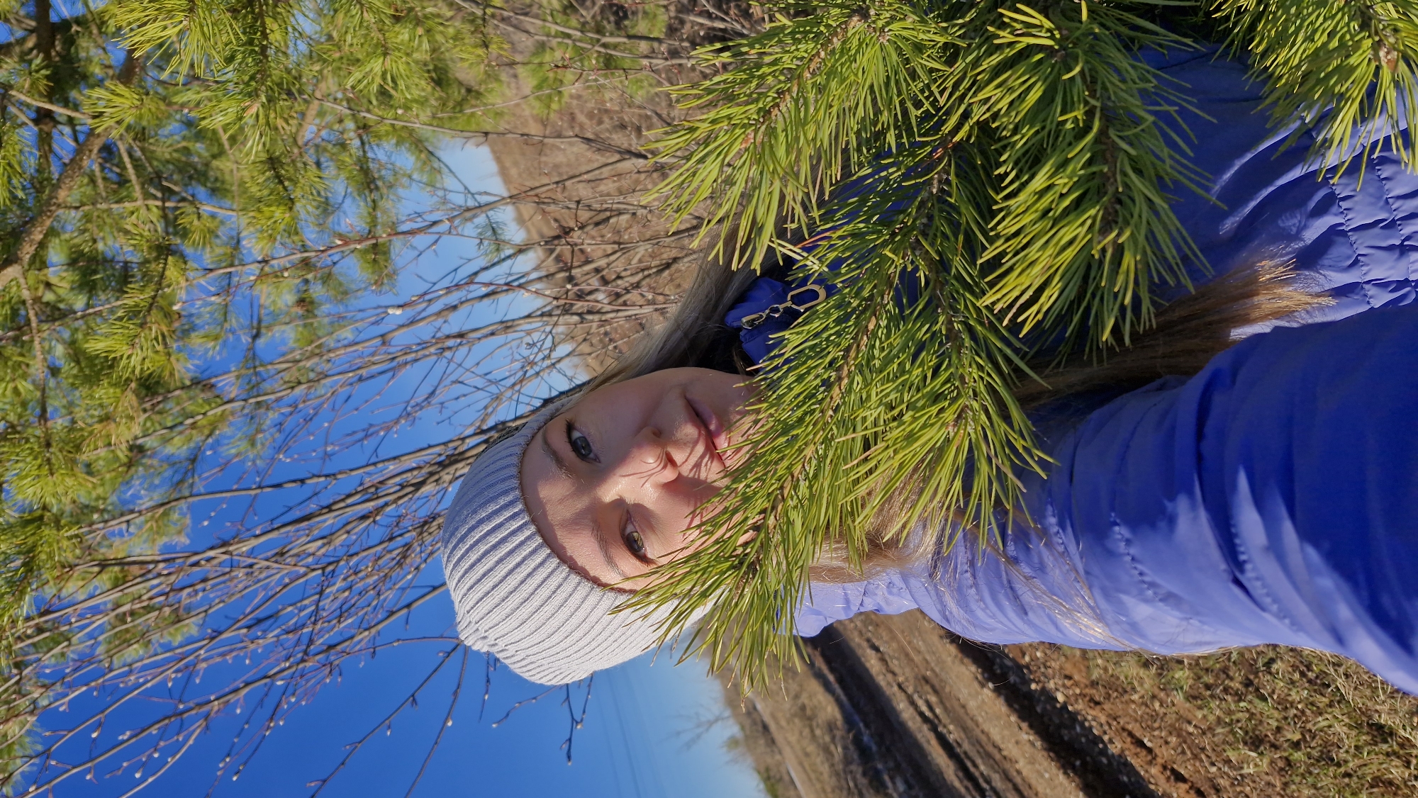 A selfie-style photograph features a young woman, possibly in her late teens to early twenties, positioned with evergreen foliage framing her face on the right. She is wearing a light gray knitted beanie and a bright blue puffy jacket. The background includes a clear blue sky and bare tree branches, indicating a winter or early spring setting. The ground appears to be a mix of dry vegetation and potentially some snow. Based on the visible elements, the location resembles a natural area or park on the outskirts of a populated area like Otchor, Russia.