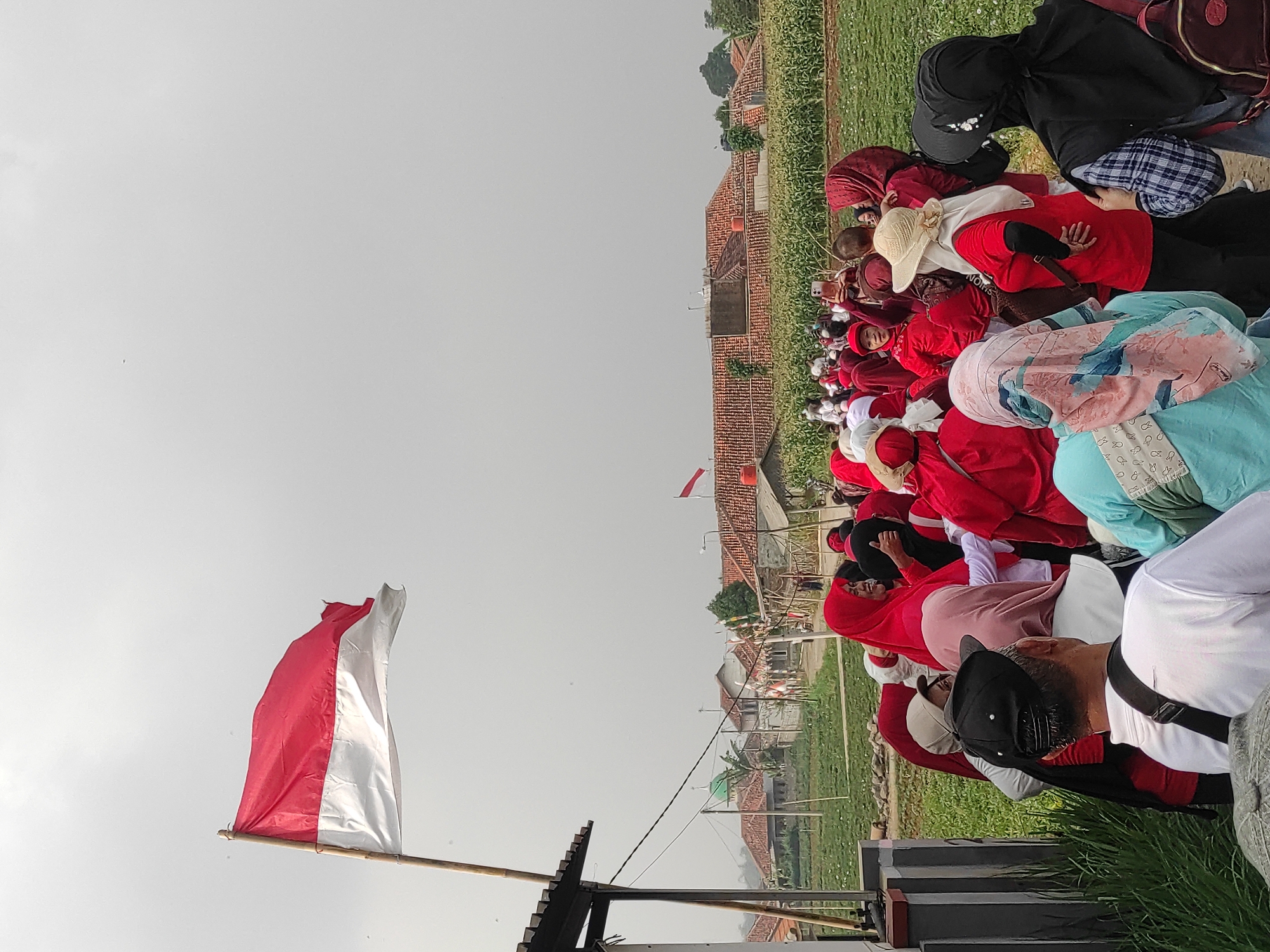 The image captures an outdoor scene in what appears to be a rural area, potentially in the vicinity of Sumedang, Indonesia, given the context. A group of people, predominantly women, are gathered, many wearing red clothing and headscarves. The Indonesian flag, a red and white bicolor, is prominently displayed, attached to a bamboo pole, and waving in the breeze. In the background, there are buildings with red tile roofs, resembling houses or small structures, situated amidst agricultural fields, possibly corn or a similar crop. The sky is overcast with a light gray hue. Some individuals are engaged in conversation, while others are looking in various directions. One person is holding up a mobile phone, seemingly taking a photograph or video. The overall atmosphere suggests a community gathering or an event taking place outdoors.