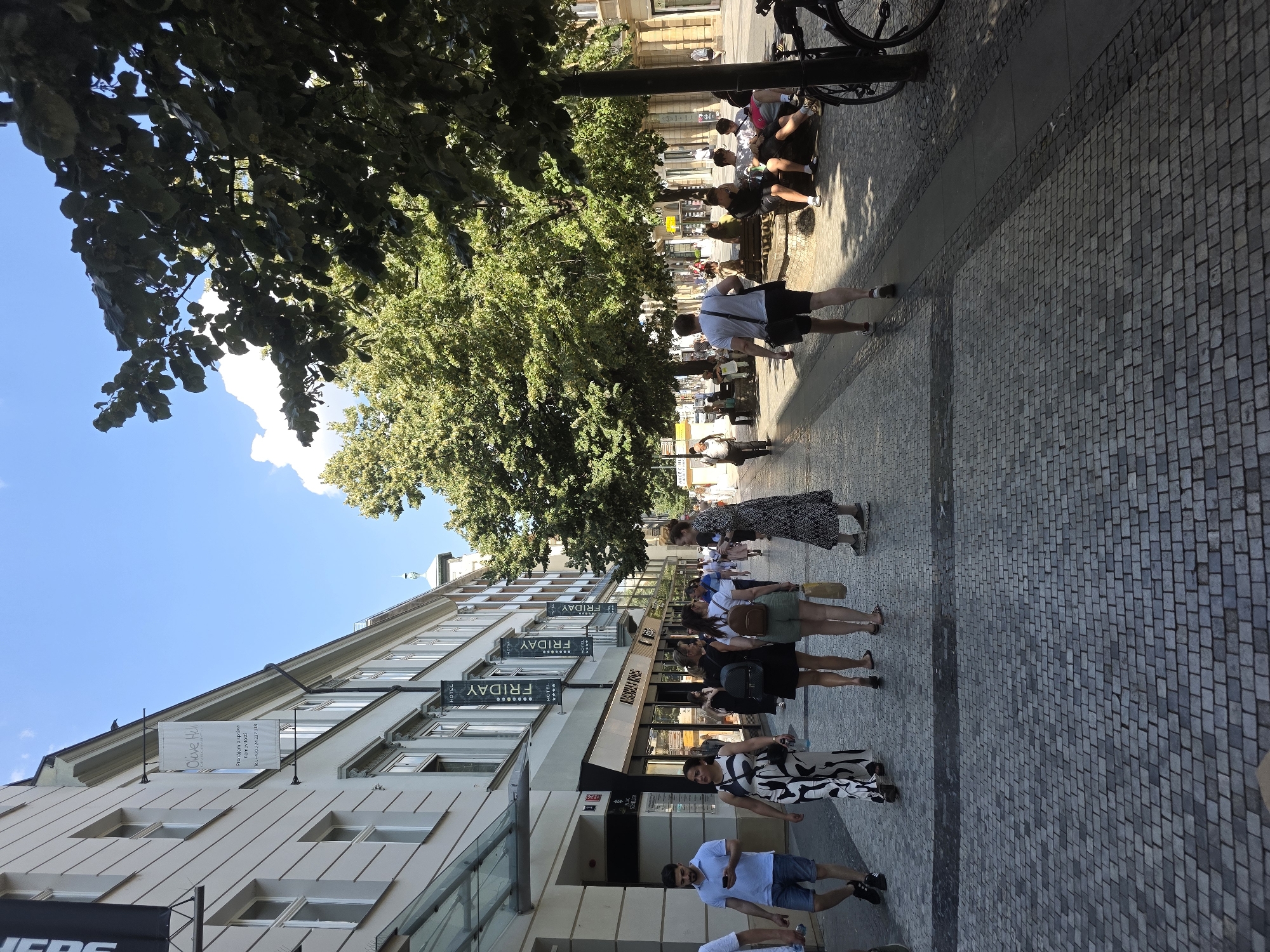 The image depicts a bustling city street scene under a clear blue sky. On the left, a modern building features a glass-covered entrance and storefronts, with banners advertising "Olive Hill" and "HOTEL FRIDAY" hanging from the facade. Several people are walking along the cobblestone sidewalk.
In the center and right of the image, a tree-lined promenade offers shade. Several individuals are seated on benches, while others walk past. A bicycle is parked near a lamppost on the right side of the frame. The overall atmosphere suggests a pleasant day in a European city, likely a commercial or tourist area given the presence of hotels and shops.