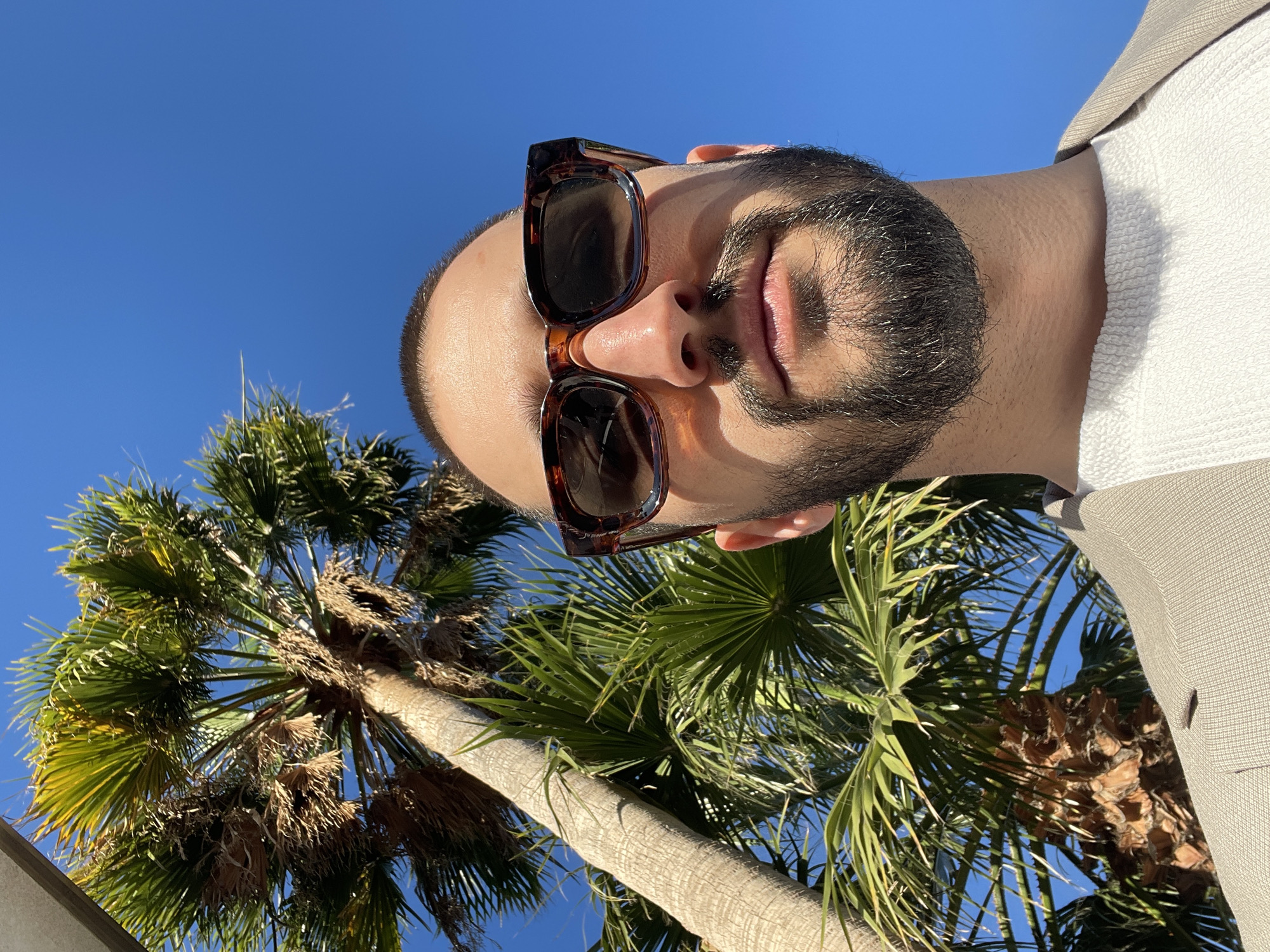 A low-angle, portrait-oriented shot captures a man looking upwards, set against a clear blue sky and lush palm trees, indicative of a sunny, subtropical environment like Santa Cruz de Tenerife. The image is taken outdoors during bright daylight, with strong sunlight creating clear shadows, and a vibrant, cloudless blue sky filling the background.
The subject is a man with a neatly trimmed beard and short hair, wearing stylish tortoiseshell sunglasses with dark lenses. His mouth is slightly open. He is dressed in a light-colored, possibly off-white or cream, textured shirt, layered beneath a light beige or tan blazer. The upward perspective highlights his head and upper torso, with his face angled towards the camera from below.
In the background, several palm trees extend upwards into the bright sky. Their green fronds are distinctly visible against the blue, with some drier, brown fronds interspersed, suggesting mature trees. The textured trunks of the palm trees are also evident. The composition evokes a relaxed, bright, and warm outdoor atmosphere. There is no visible text within the image.