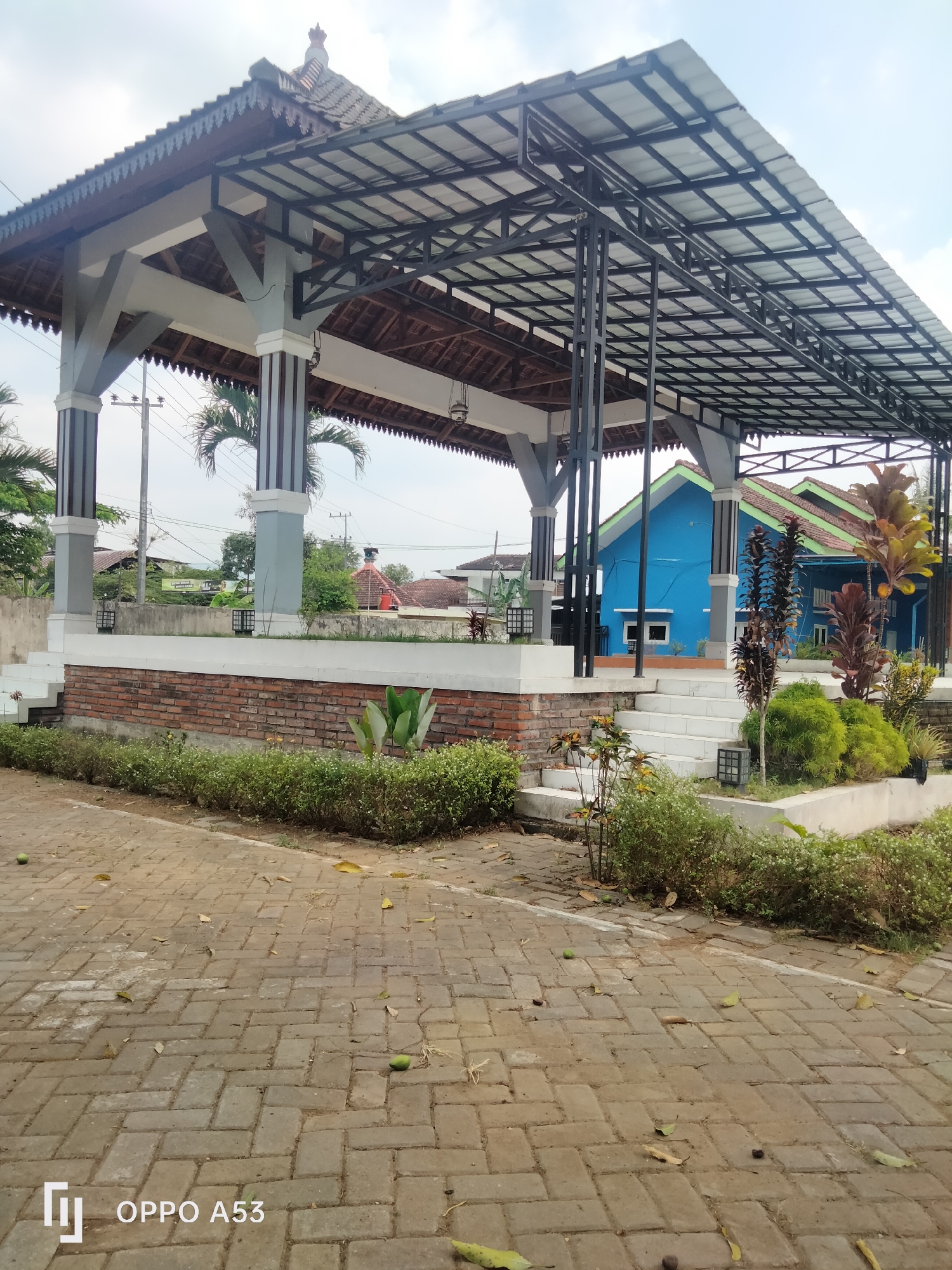 The image shows an open-air pavilion with a gray and white structure featuring a tiled roof and a metal frame. The pavilion is elevated on a brick base and has white stairs leading up to it. In the background, there are two blue houses with green roofs. The ground in front of the pavilion is paved with interlocking bricks. Various plants and bushes are situated around the pavilion and its base. The scene appears to be in a semi-rural or residential area, possibly a public space within Karangan, Indonesia. The sky is visible with some clouds.