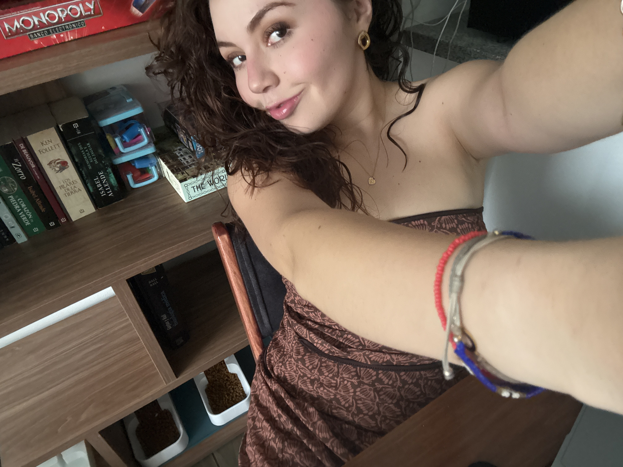 A selfie captures a young woman with curly brown hair, wearing gold hoop earrings and a delicate gold necklace. She's dressed in a strapless brown and beige patterned garment. The background includes a wooden shelf stacked with items. On the top shelf, a "MONOPOLY" board game box is visible along with various books, including titles by Isabel Allende and Ken Follett. A "The World" game box is also present. Below, on the lower shelves, there is an assortment of books, including medical texts, and white trays containing brown granular material. The location appears to be indoors in Mexico City, Mexico, as indicated by the presence of books by Spanish-speaking authors and the Spanish translation of "MONOPOLY."