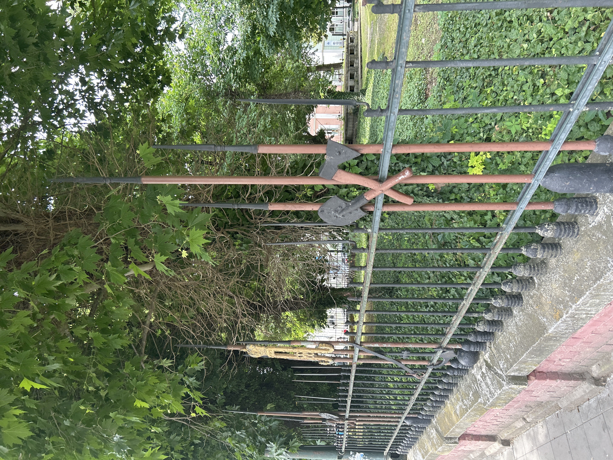 The image displays a detailed metal fence running horizontally across the midground, suggesting a boundary for a park or historical site. The fence's vertical pickets are elaborately designed, featuring decorative finials that resemble historical weaponry and tools such as spears, axes, and shovels, rendered in dark grey, black, and rusty brown tones, indicative of weathered metal or specialized paint.
Behind this distinctive fence, a lush green environment unfolds, filled with dense trees showcasing vibrant foliage and ground covered with ivy. Through the gaps in the greenery, faint views of distant buildings are visible, including one light-colored structure and another with a reddish-pink hue. In the immediate foreground, below the fence, a concrete or stone base is apparent, followed by a border of red bricks and a grey paved sidewalk.
No specific activity or event is taking place; the scene is static and serene. The bright, natural light filtering through the tree canopy indicates daytime, likely a sunny or partly cloudy day. The unique, armament-themed design of the fence strongly suggests a historical monument or a memorial location, a detail consistent with the provided context of Pleven, Bulgaria, a city known for its historical significance. No text is visible in the image. The overall impression is one of tranquility and historical reflection.