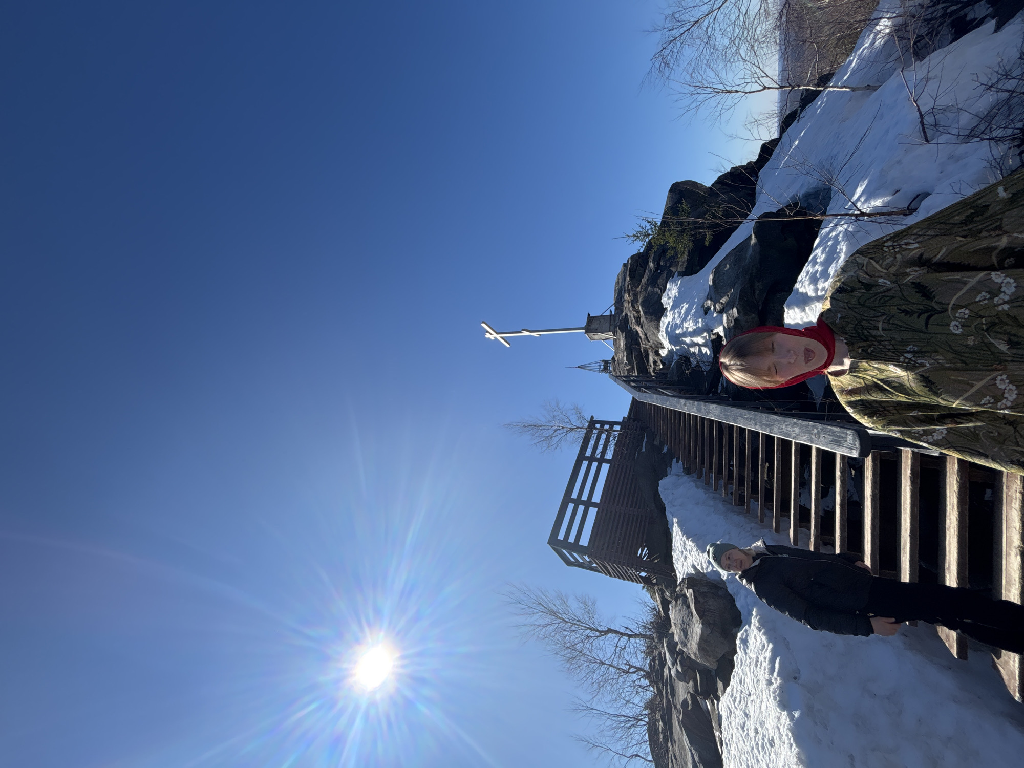 Two individuals are positioned on a wooden stairway ascending a rocky, snow-covered hillside in Gubakha, Russia. Above them stands a prominent Orthodox cross. The sky is clear and bright. The sun is shining to the bottom left of the photo. The stairway features wooden planks and railings. Rocks and snow are visible around the stairway. One individual is dressed in a camouflage style jacket and a red hood. The second individual is wearing a black jacket and dark trousers, and a knit cap.