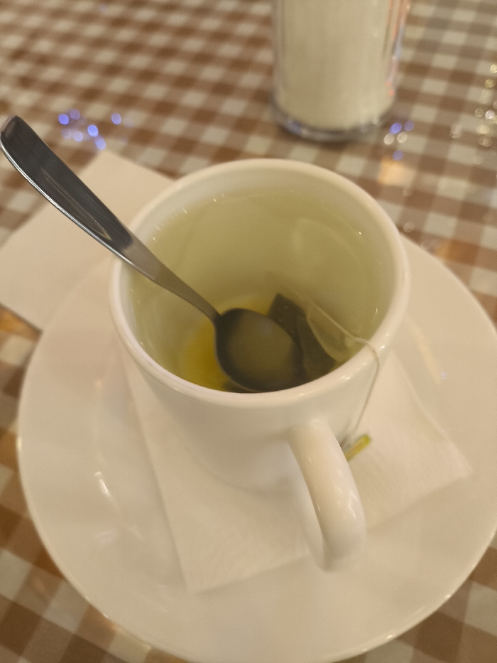 A close-up shot captures a white teacup on a matching saucer. Inside the cup, a tea bag and a silver spoon rest in a small amount of liquid. A folded white napkin cushions the cup. The saucer's surface is smooth and pristine. The scene is set against a checkered tablecloth, adding a textured background. A partially visible container, possibly filled with sugar, stands in the upper right corner, slightly out of focus.