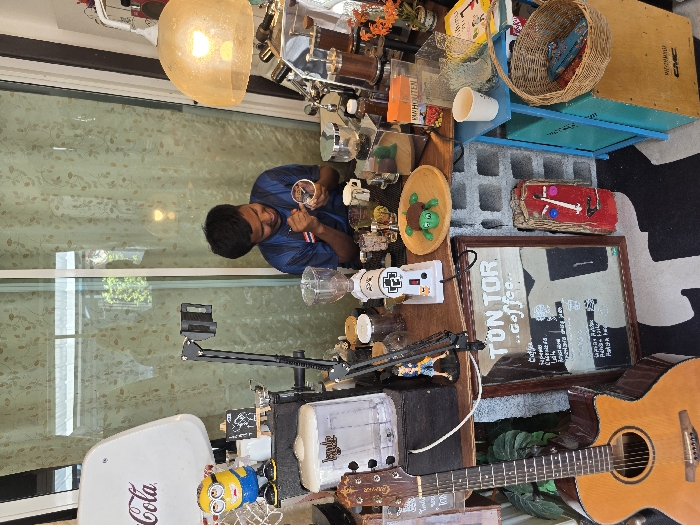A man in a blue shirt stands behind a counter with various items, including coffee-making equipment, small figures, and decorative objects. A guitar lies on the right edge of the image, with a vintage Coca-Cola sign and a Minion toy partially visible on the left. A framed menu leaning against the guitar displays the words "TON TOR...coffee". A basket sits on a blue shelf in the background.