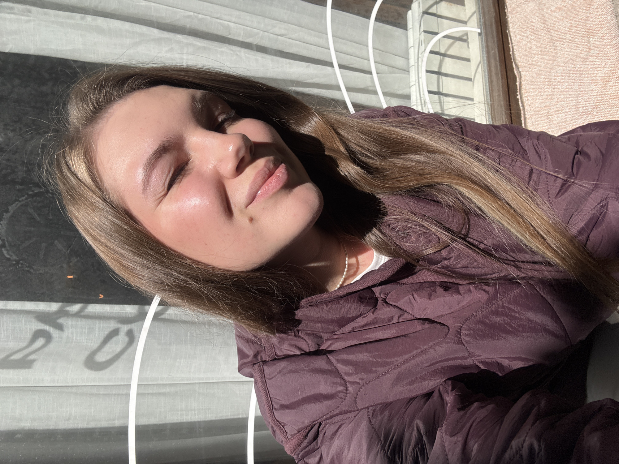 A young woman with light brown hair and a maroon puffer jacket poses in front of a window with white vertical blinds and a peach-colored patterned wall, likely inside a building. She has her eyes closed and is smiling slightly, with sunlight illuminating her face. A white pearl necklace is visible at her neck. Based on available information, it is not possible to definitively determine the location as the city of Belgrade, Serbia. The picture focuses on the woman and her immediate surroundings, lacking specific details that would pinpoint the exact location.