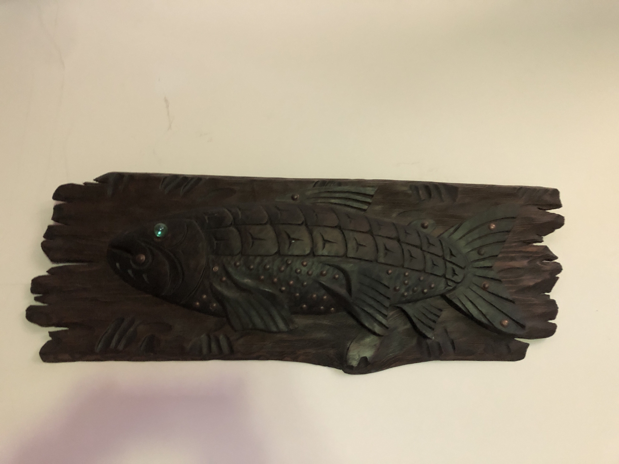A carved wooden plaque of a fish hangs on a plain, light-colored wall. The plaque is rectangular with rough, uneven edges, resembling driftwood. The fish is depicted in profile, facing left, with detailed scales and fins. It has a large eye, which appears to be a green gem. Small, round, copper-colored studs are embedded across the body of the fish, possibly representing spots. The wood is dark brown and appears to be stained or aged. The plaque is positioned horizontally on the wall. There is no indication of the city of Oryol, Russia, within the image itself.